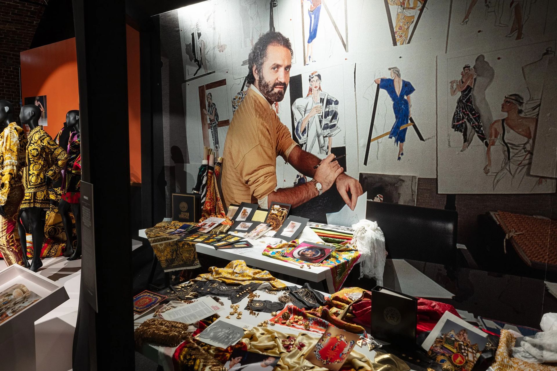 A museum exhibit displays colourful fabrics and accessories on a table in front of a large wall photo of a designer at work, evoking the vibrant atmosphere of fashion exhibitions, with sketches and mannequins in ornate garments in the background.
