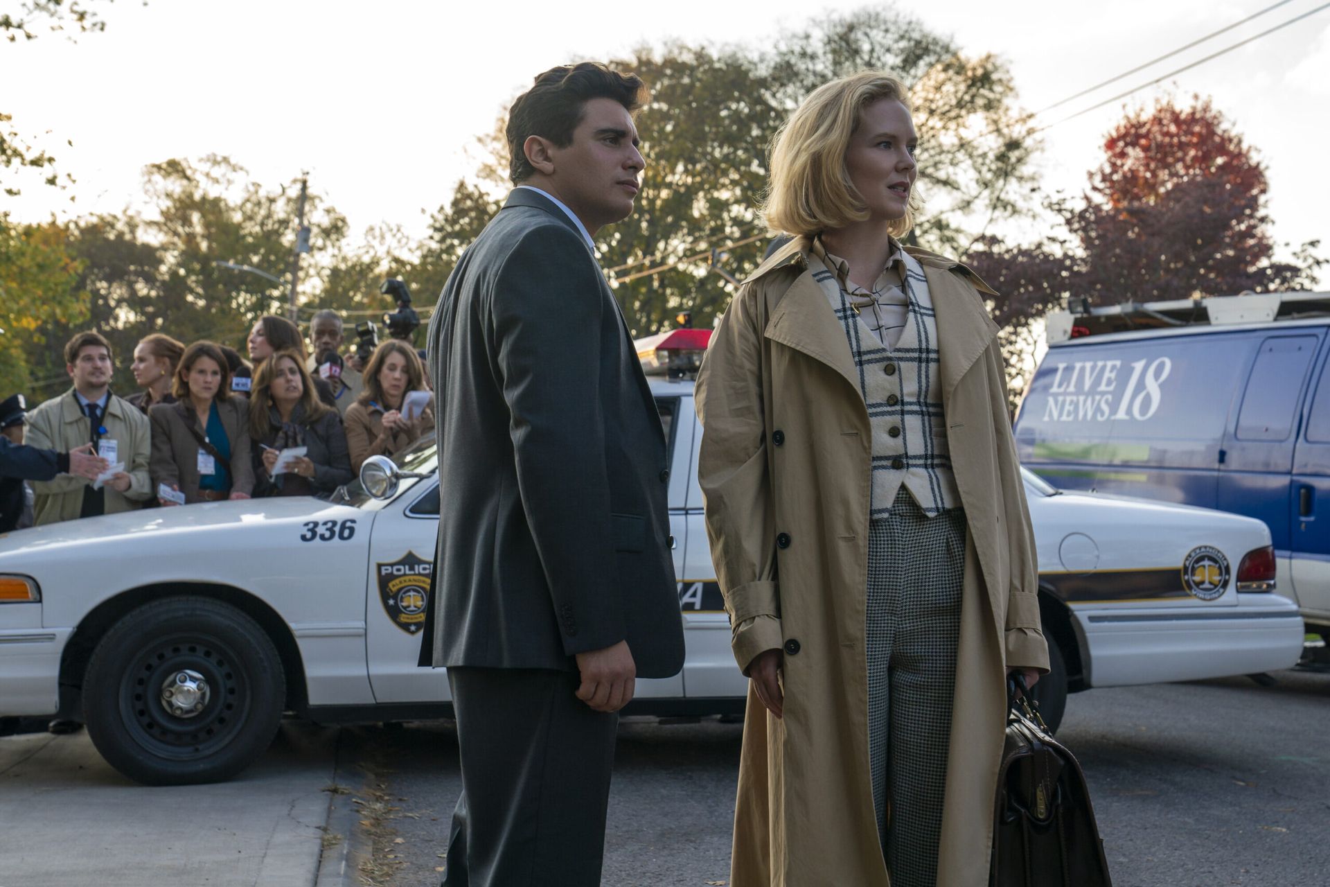 A man in a suit and a woman in a checked shirt and long trench coat stand before a police car, evoking classic Murder Thriller vibes. Journalists and a news van linger nearby on the suburban street—just like scenes from Scarpetta starring Nicole Kidman.