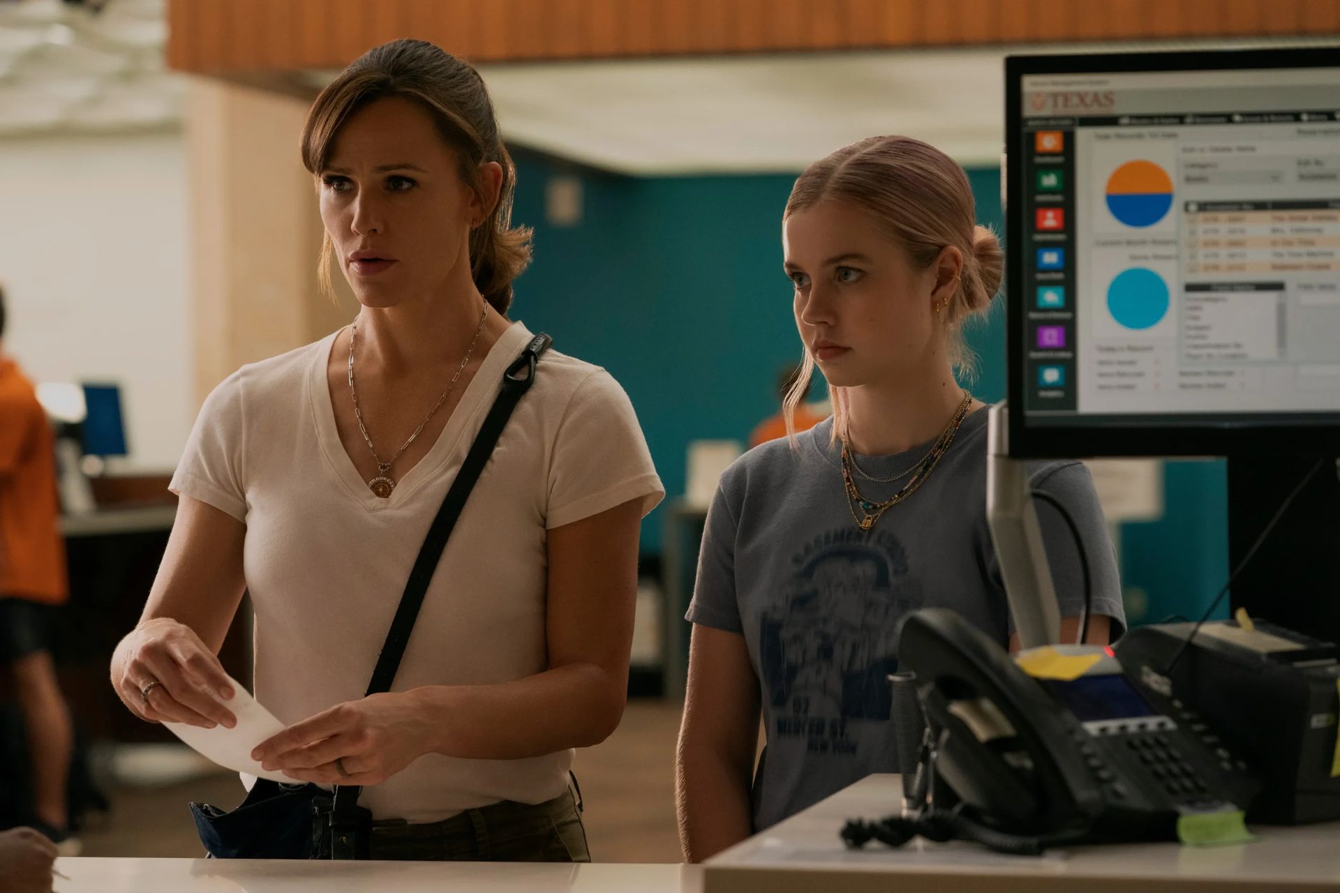 Two women stand at a counter in an office setting, one holding a piece of paper with a concerned look. The scene evokes the tension of The Last Thing He Told Me Series 2, with a computer monitor and office supplies on the counter.