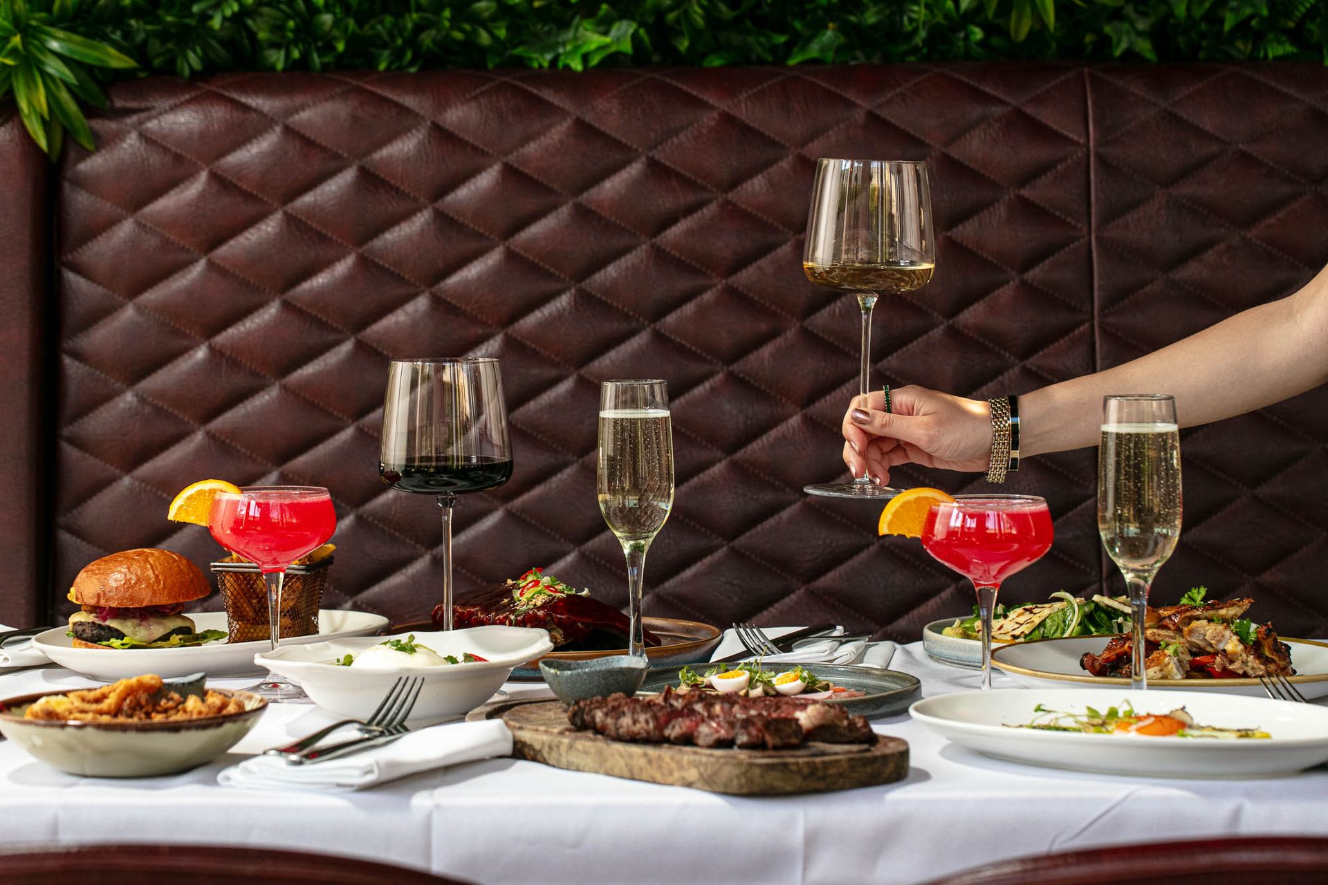 A table set with various dishes—steak, salads, burgers, and colourful cocktails—is perfect for a weekend guide. Three wine glasses and two champagne flutes are visible as a hand raises a glass for a toast against a tufted leather banquette background.