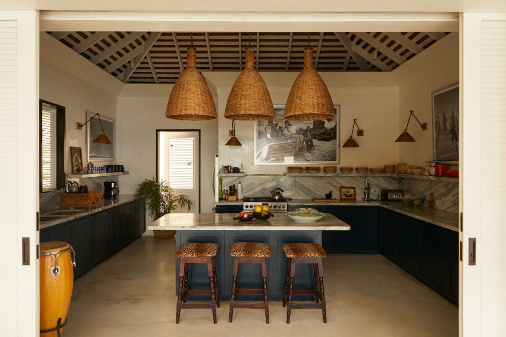 Spacious kitchen with dark cupboards, marble worktops, a large island with stools, woven pendant lights, and tropical decor. Sunlight streams through windows, highlighting plants and wall art—a signature look from Barlow & Barlow.