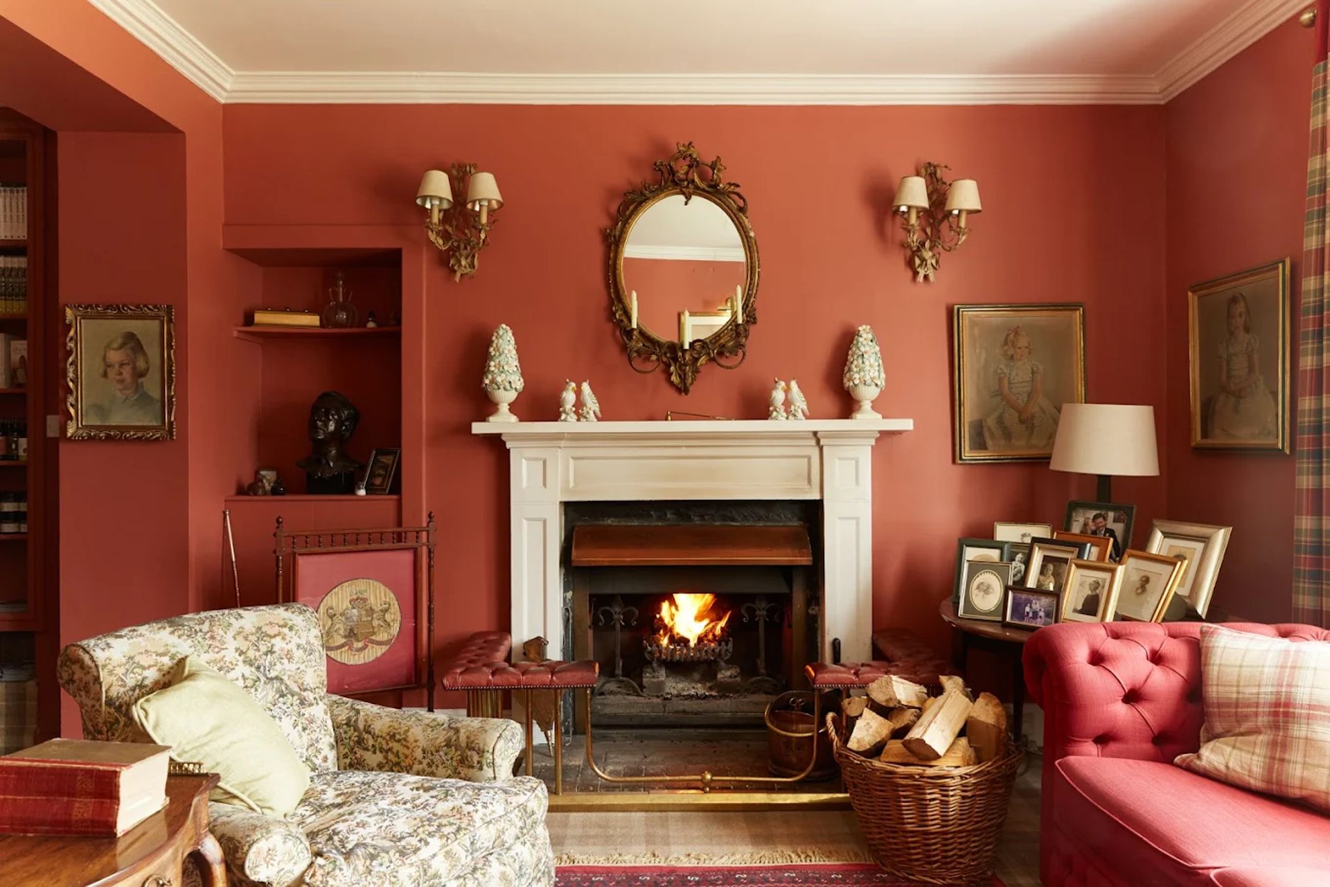 A cosy living room reminiscent of Kinloch Lodge, with red walls, a lit fireplace, vintage furniture, framed portraits, a round mirror above the mantelpiece, and a basket of firewood beside a red sofa and floral armchair.