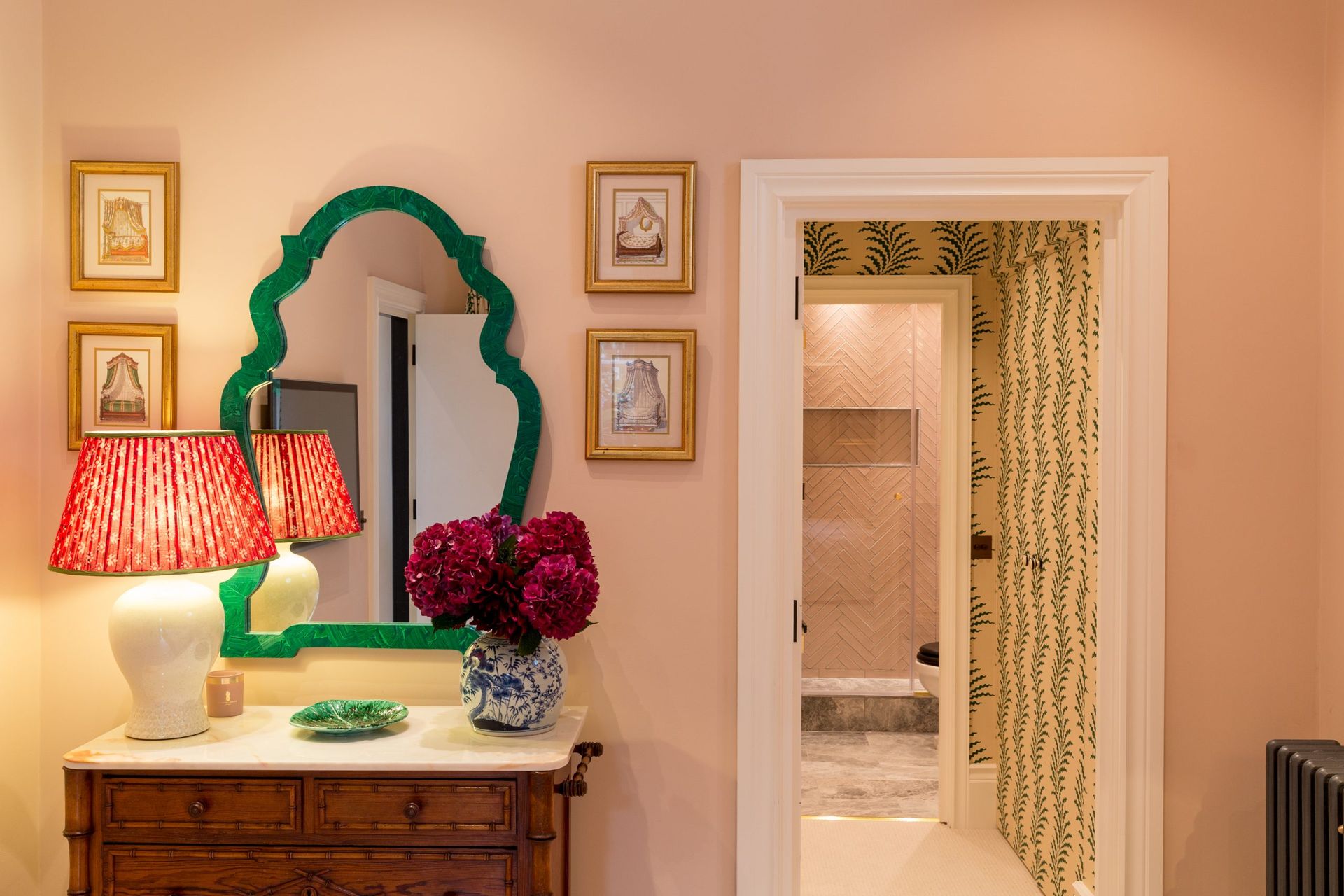 A warmly lit room with a vintage wooden chest of drawers, a white lamp with a red patterned shade, and a green-framed mirror captures the Interiors Insider style of Lucy Barlow; an open door reveals a wallpapered hallway and bathroom beyond.