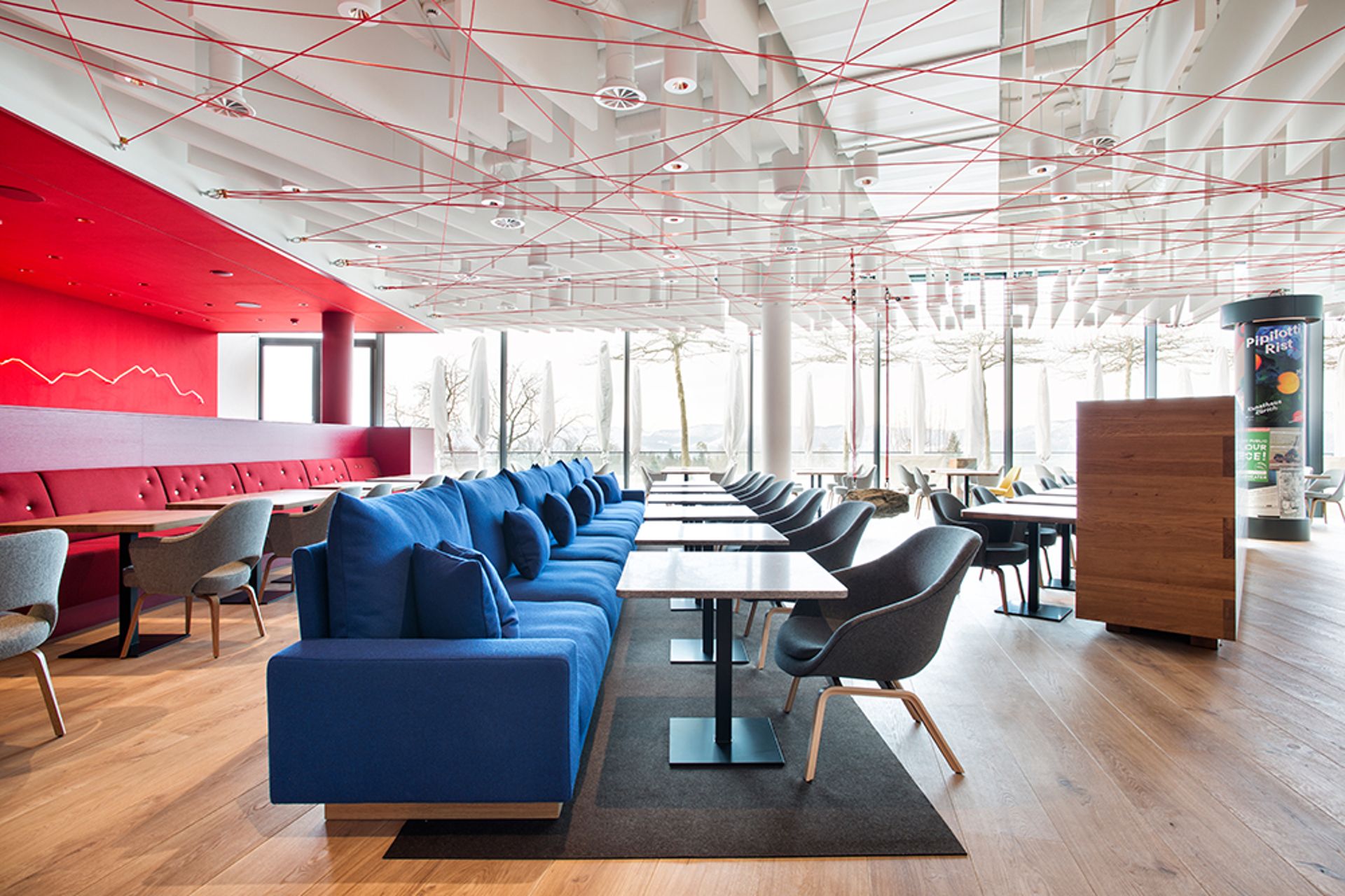 Modern, spacious lounge at the Dolder Grand in Zurich, featuring blue sofas, grey chairs, and wooden tables on a light wood floor. Red walls and ceiling have criss-crossing lines, whilst large windows fill this fairytale hotel space with natural light.
