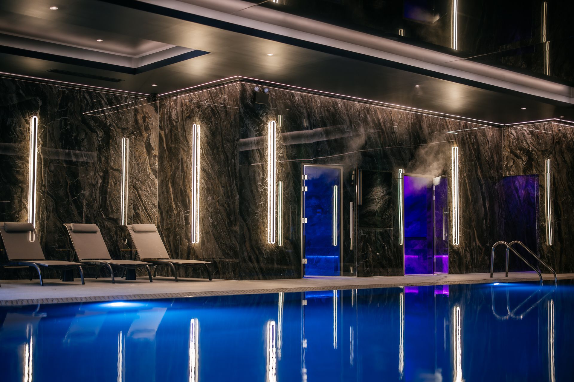 Indoor swimming pool with blue water, modern lounge chairs, and illuminated vertical lights on dark marble walls. A glass steam room door glows with purple light in the background, creating a luxurious, relaxing ambience—making this retreat one of the best spas outside London.