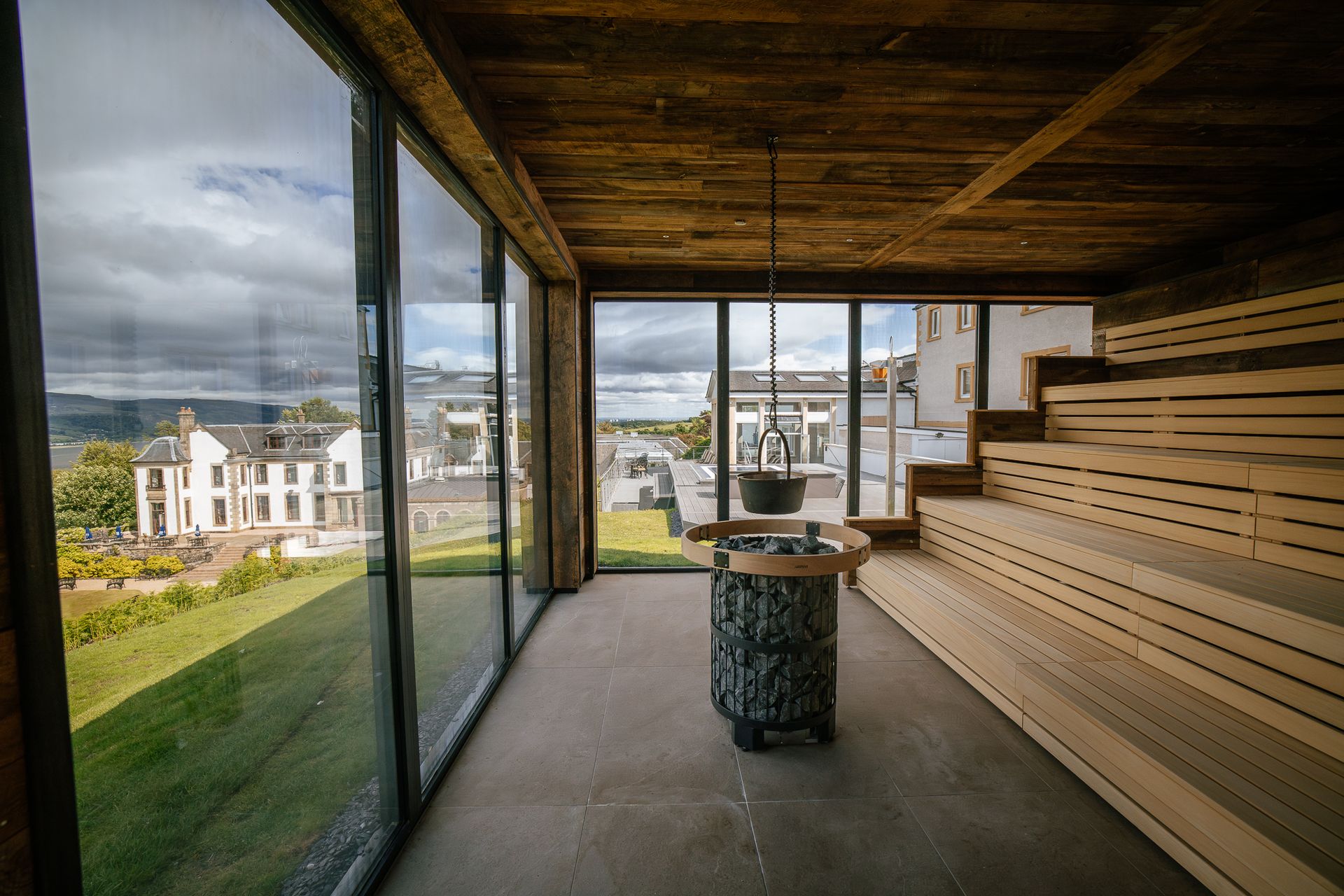 Modern wooden sauna with large glass windows overlooking a scenic, green outdoor landscape and white buildings under a cloudy sky—an inviting retreat that rivals some of the best spas outside London.