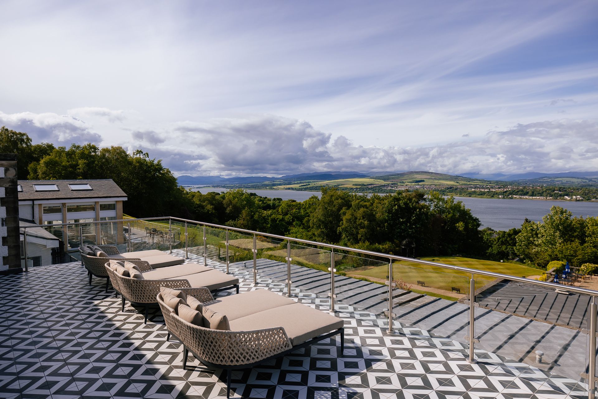 Modern patio with patterned tiled floor, lounge chairs, and glass balustrade overlooks green hills, trees, and a large lake under a partly cloudy sky—a tranquil setting reminiscent of the best spas outside London.