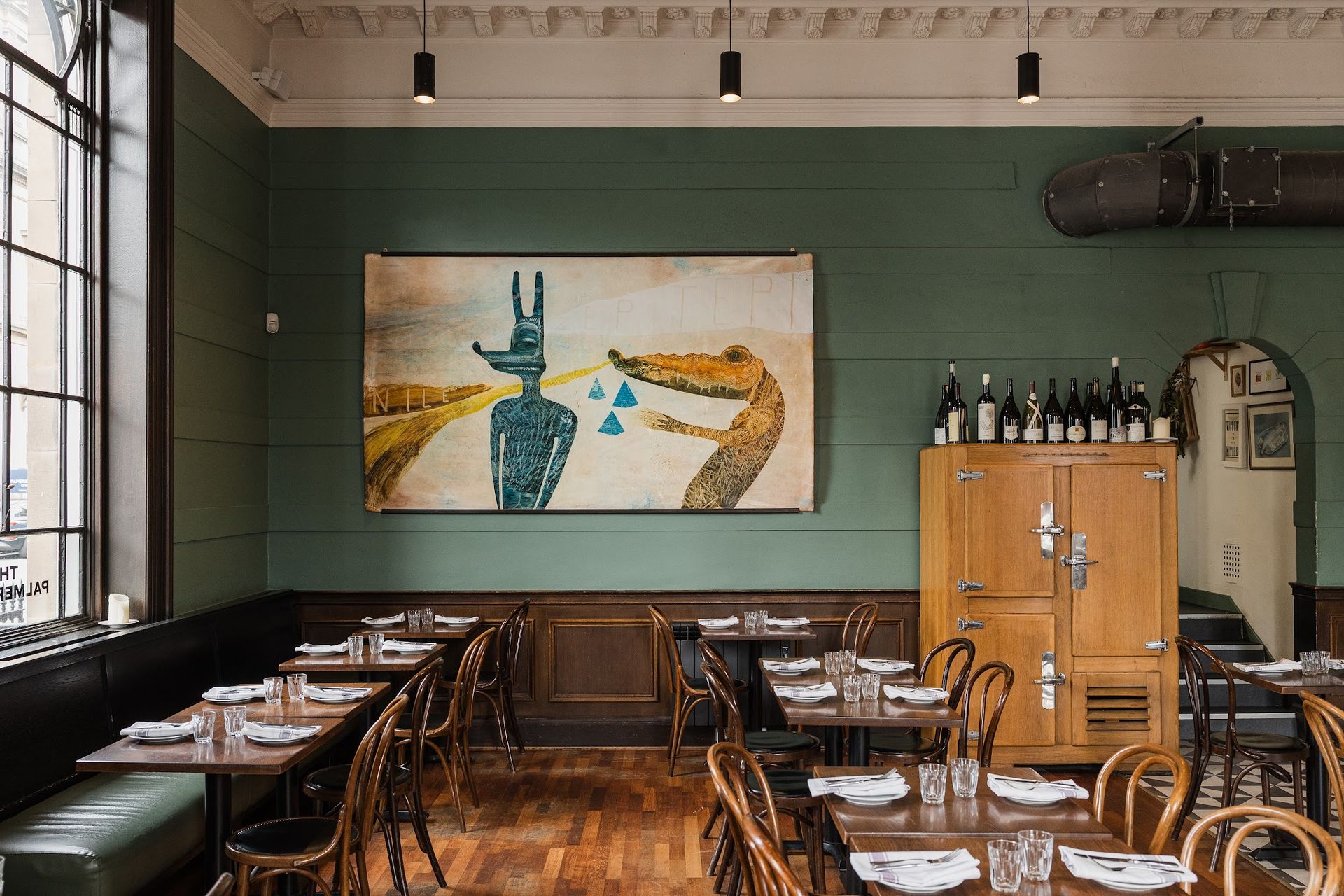A cosy restaurant in Edinburgh with green walls, wooden tables and chairs, a large painting of abstract animals, wine bottles on a shelf, and a vintage fridge. Natural light streams in from tall windows—an inviting spot that’s often recommended in any true Insider’s Guide to Edinburgh travel.