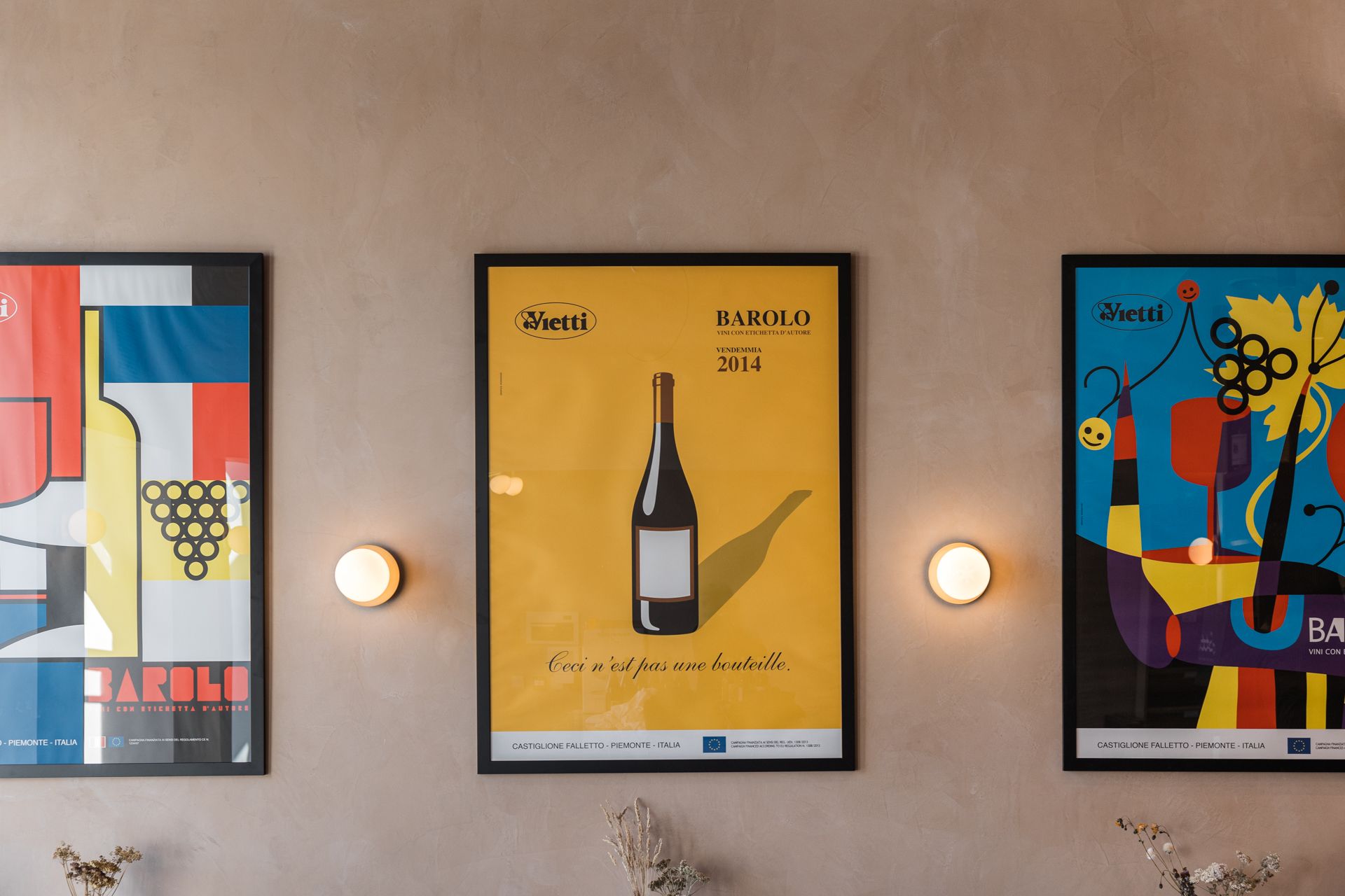 Three colourful framed posters hang on a beige wall, each featuring wine-themed art reminiscent of an insider’s guide to Edinburgh’s vibrant bar scene. Two small round wall lights are positioned between the posters, softly illuminating the space.