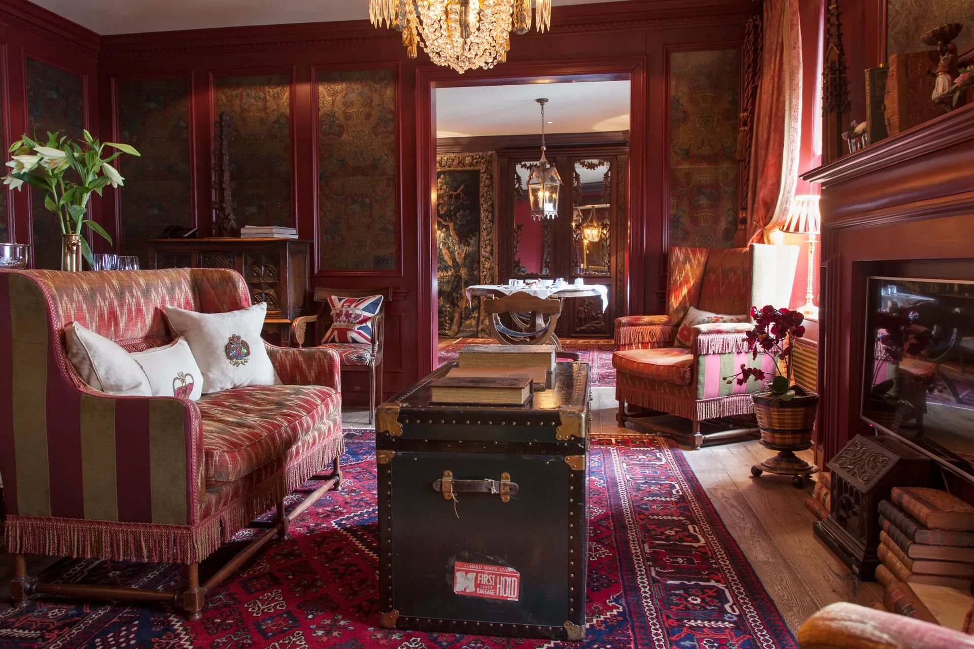A vintage living room in Edinburgh with red walls, patterned rugs, a striped sofa, antique chairs, a trunk used as a coffee table, fireplace, chandelier, and plants. Like an Insider’s Guide to classic interiors, the space extends into a dining room visible through an open doorway.