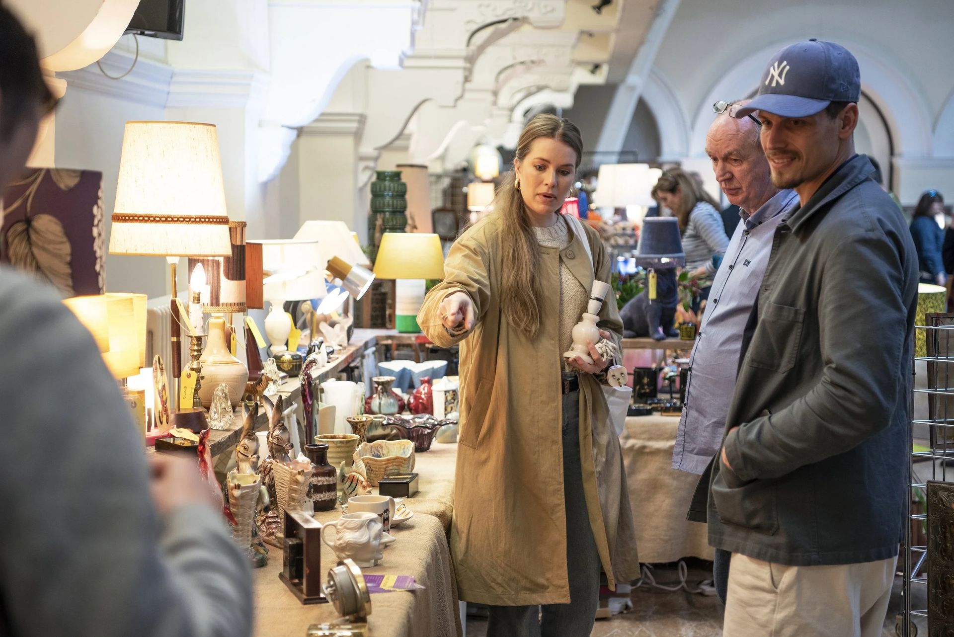 At an indoor market inspired by London flea markets, a woman in a tan coat acts as a guide, pointing at decorative objects and lamps on the table, whilst two men—one in a blue baseball cap—stand nearby observing.