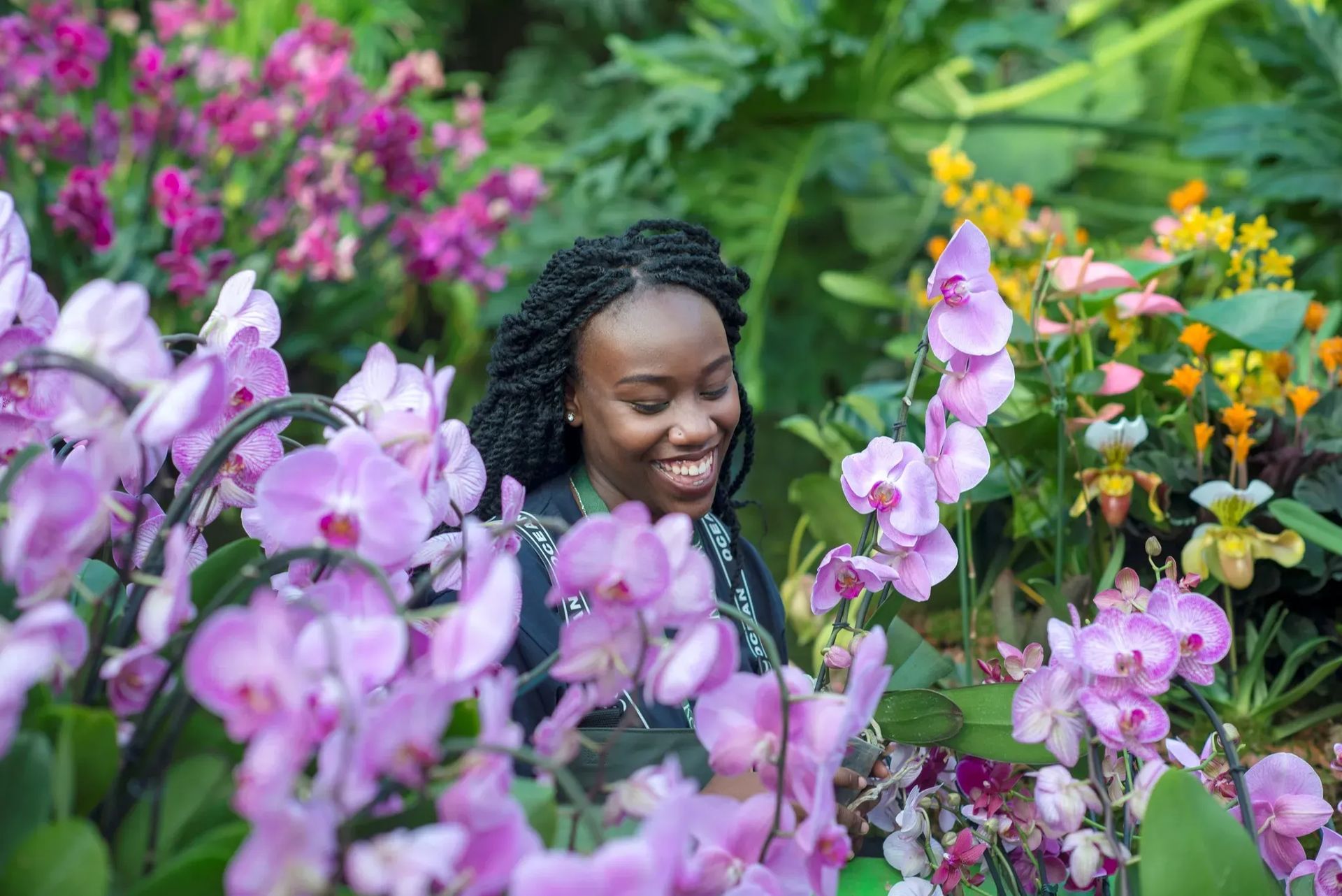 A woman smiles joyfully whilst surrounded by vibrant pink, purple, and yellow orchids and lush green foliage in a garden inspired by London February pop-ups.