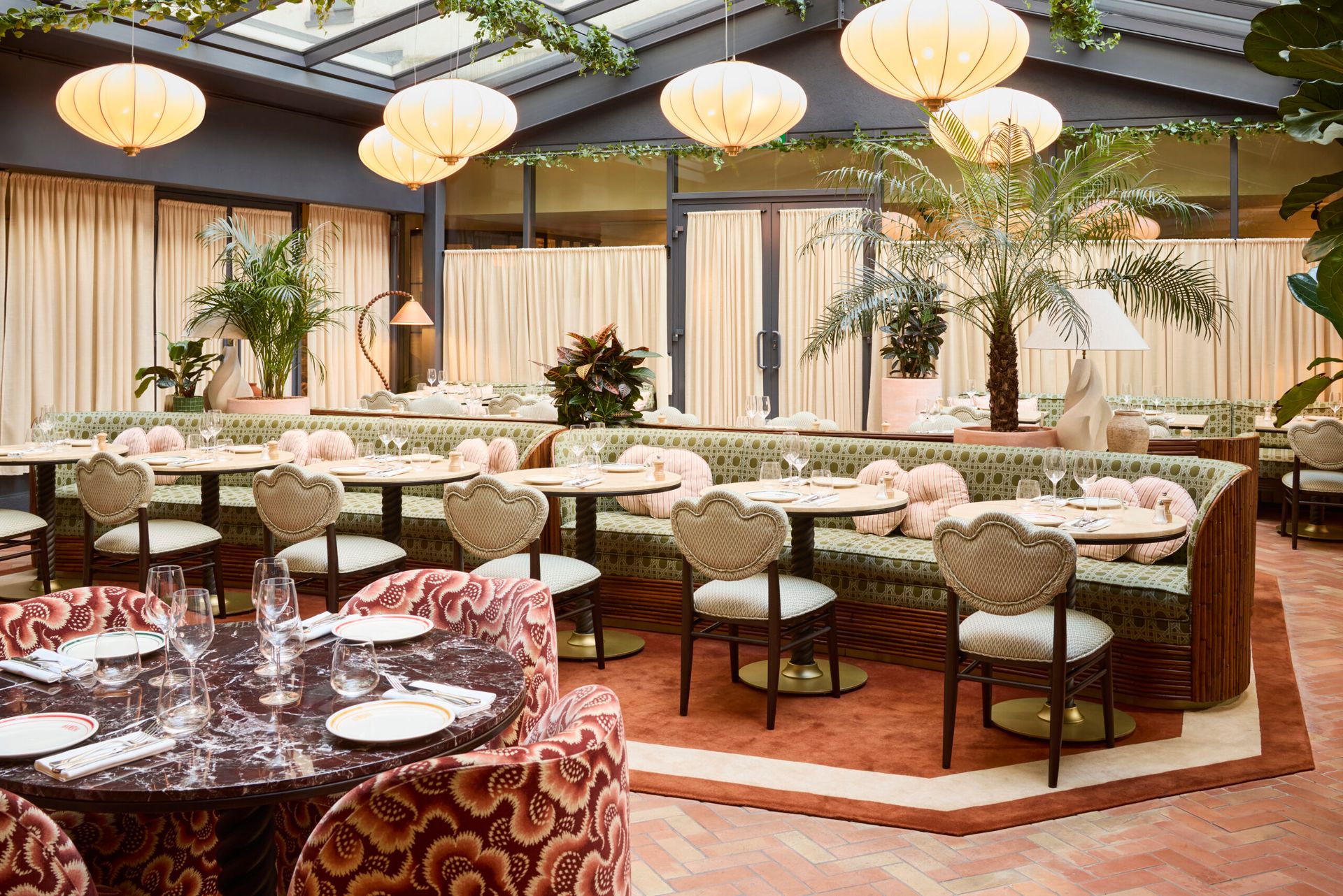 A stylish restaurant dining area at The Hoxton features patterned chairs, marble tables, upholstered banquettes, large round light fittings, indoor plants, and natural light from a glass ceiling. The decor showcases soft pastel tones and elegant details.