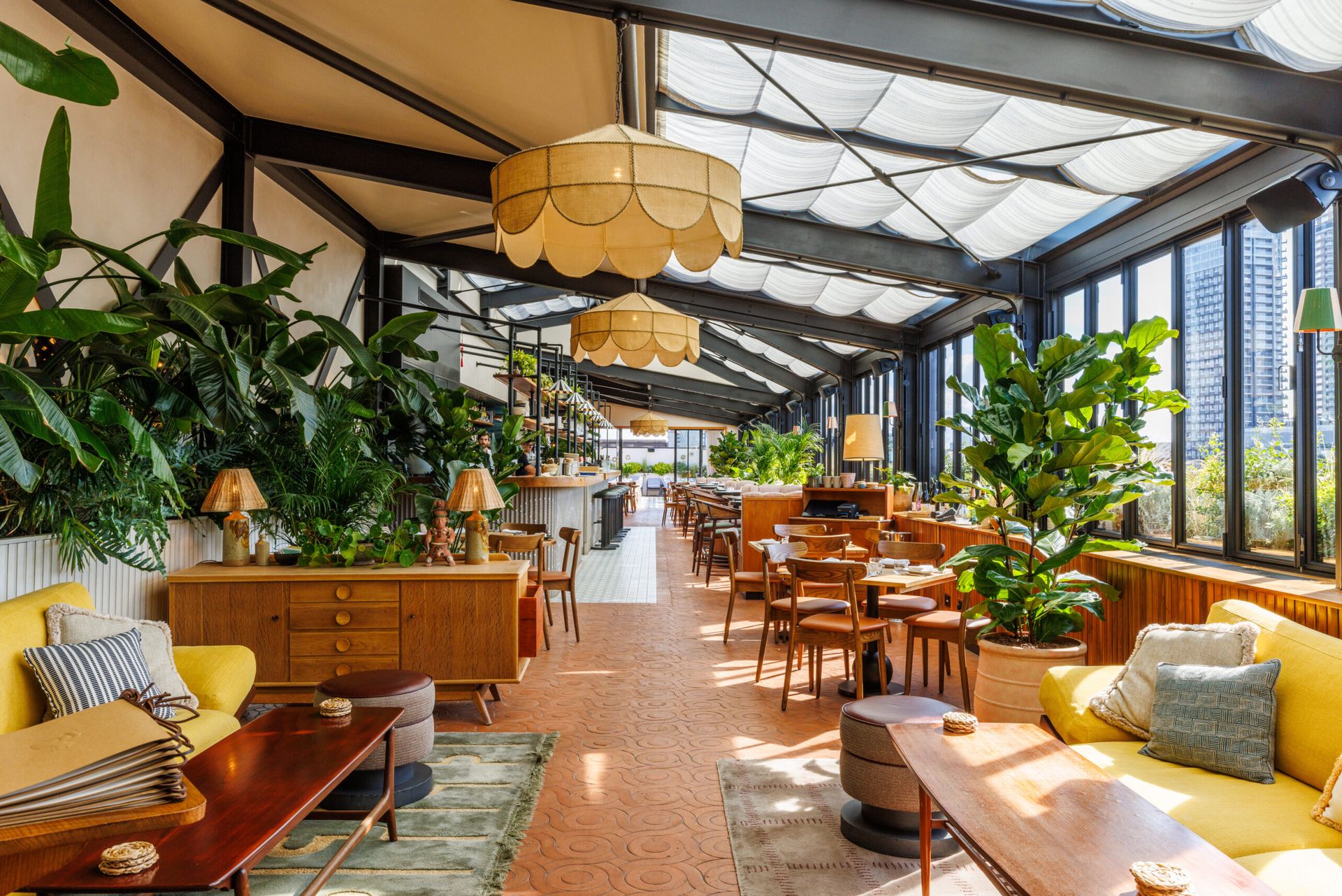 A bright, stylish restaurant at The Hoxton with large windows, indoor plants, wooden tables and chairs, cosy sofas, and unique hanging lights. The decor mixes vintage and modern elements for a warm, inviting atmosphere.