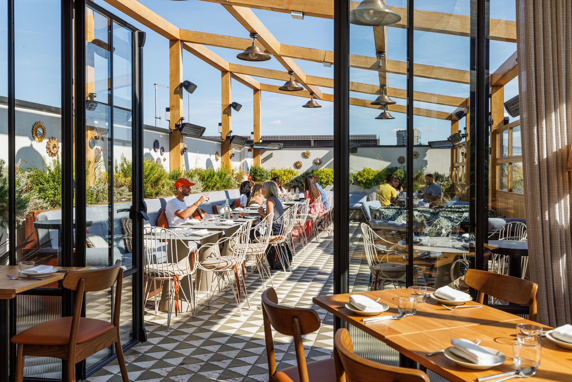 A sunlit restaurant patio at The Hoxton with glass walls and a wooden pergola, where diners enjoy meals. The space features patterned tile flooring, lush greenery, and modern décor.