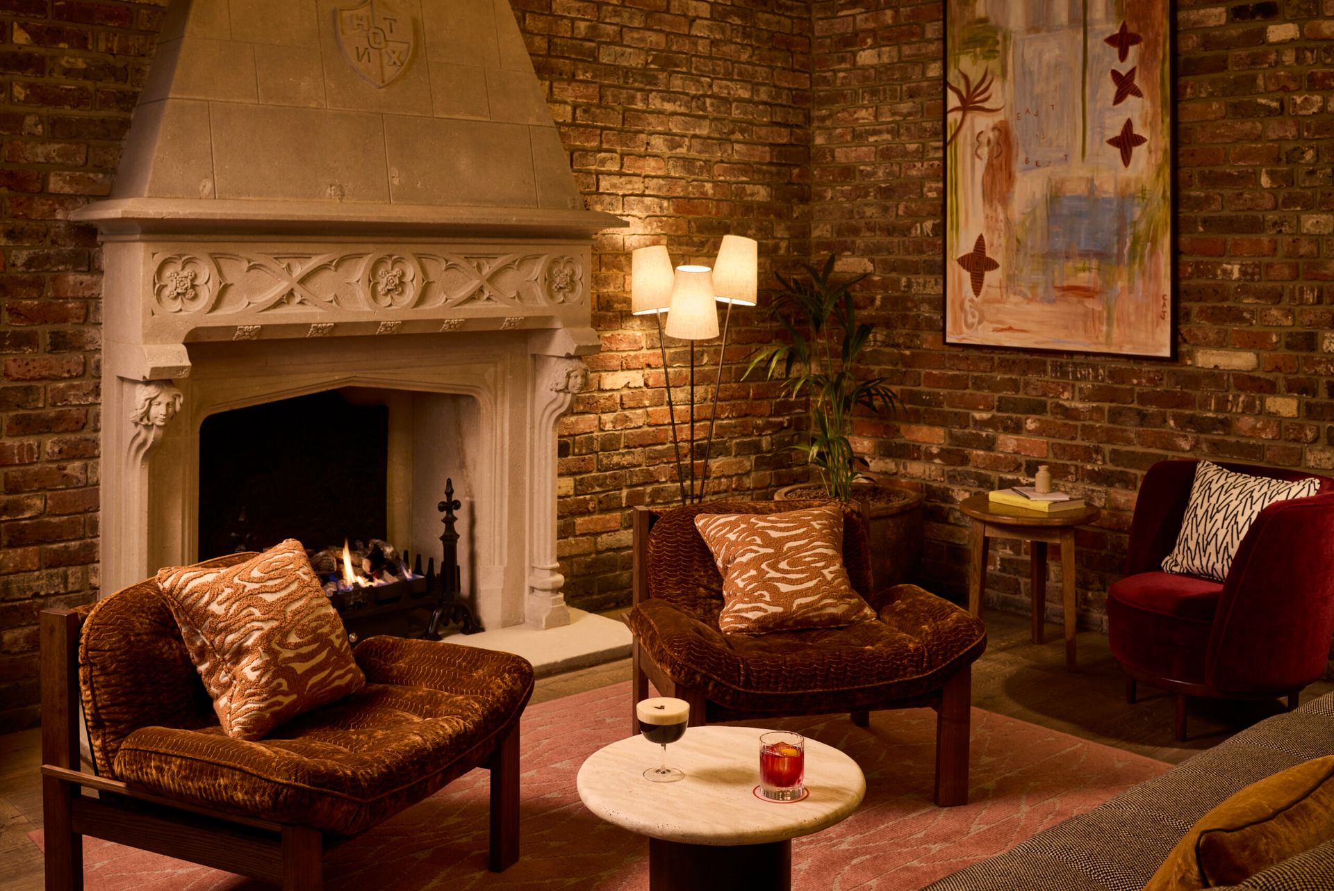 A cosy living room inspired by The Hoxton, with a lit fireplace, brick walls, two brown cushioned chairs, a small round table, a floor lamp, a red armchair, and a modern painting hanging on the wall.