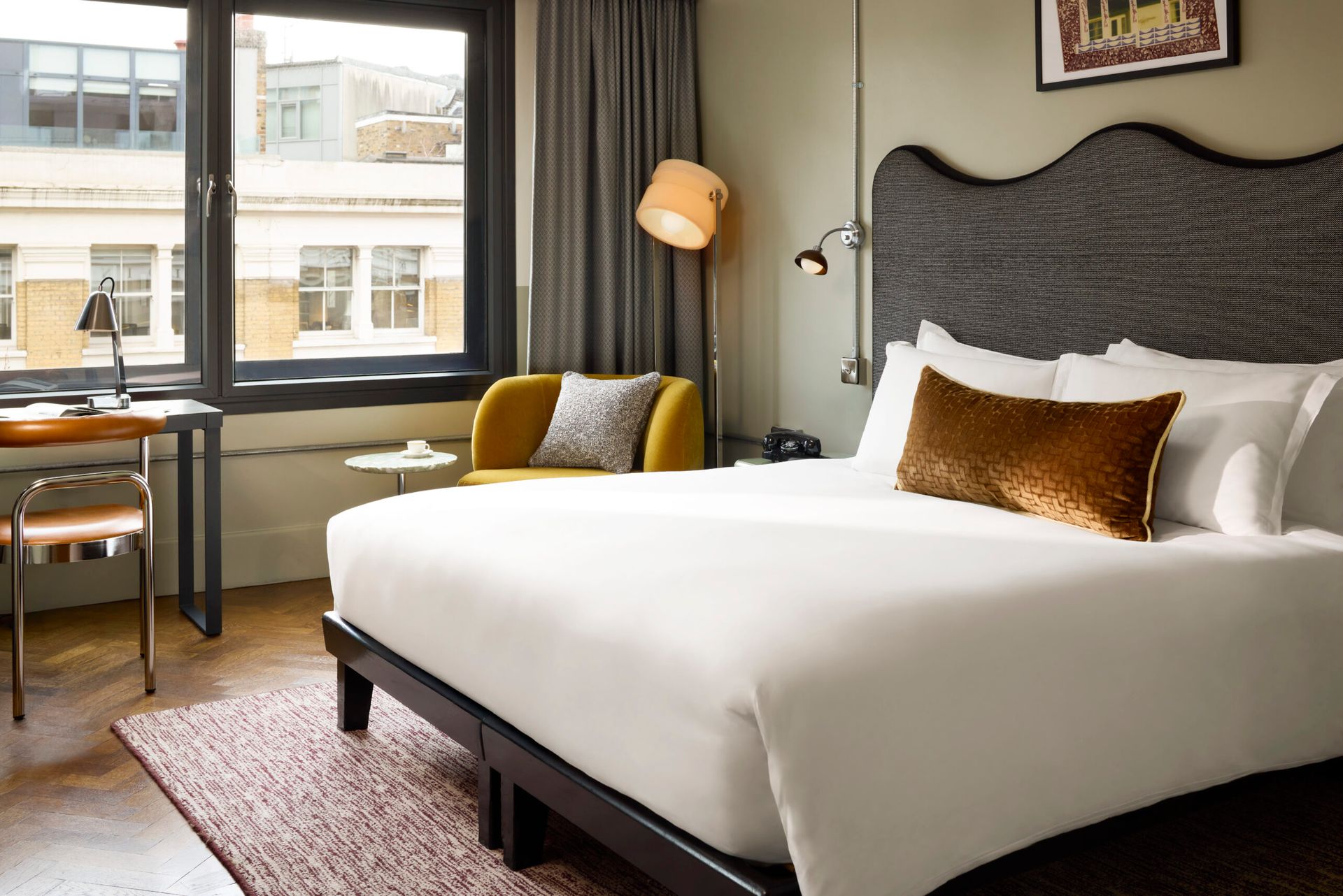 A modern hotel room at Hoxton Shoreditch in London features a large bed with white linen and a brown accent cushion, a yellow armchair, a desk by the window, standard lamp, and framed art. Natural light fills the stylish space.