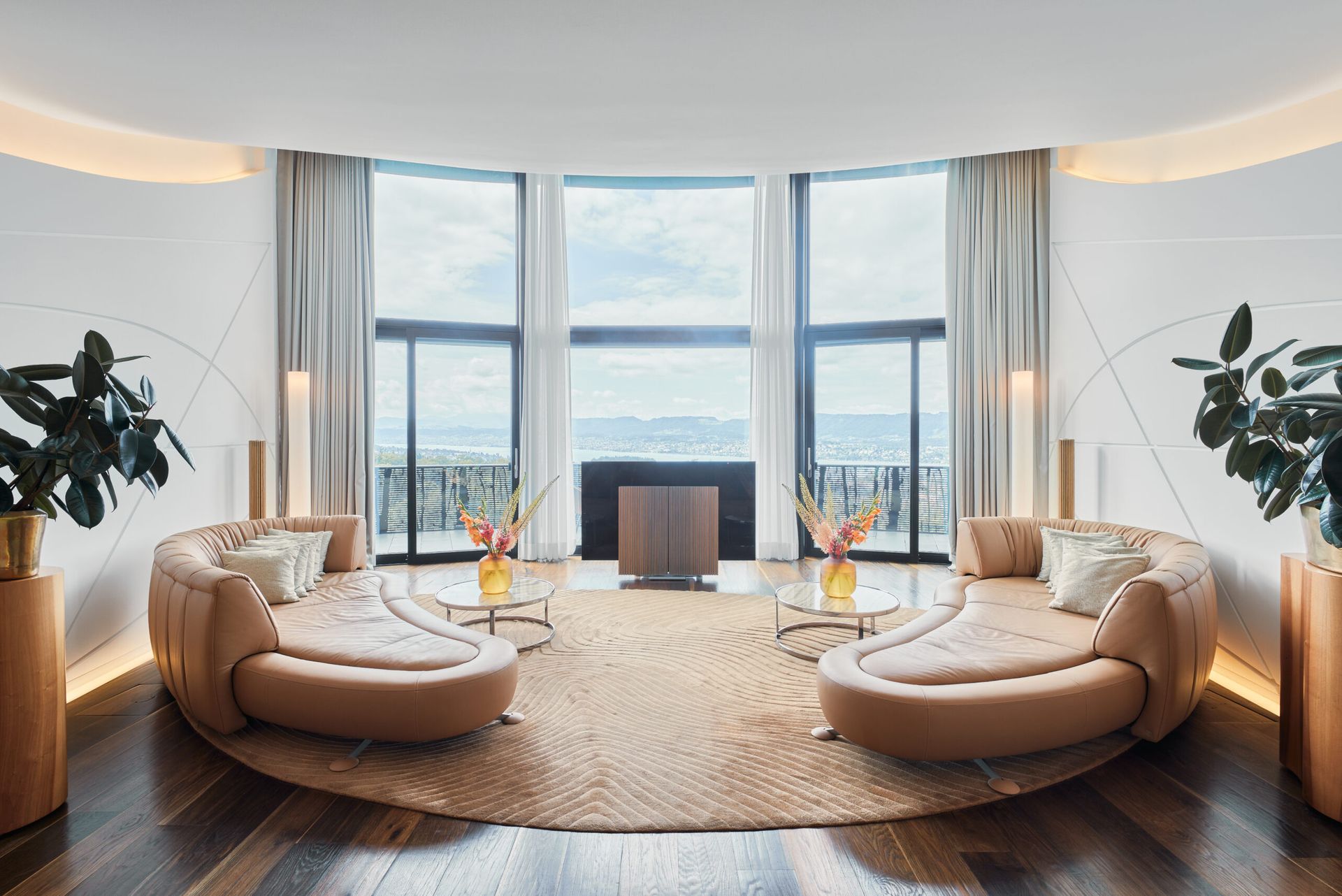 A modern, elegant living room inspired by the Dolder Grand in Zurich, with two curved tan sofas, glass coffee tables adorned with flowers, large windows overlooking a scenic view, and abundant natural light highlighting wooden floors.