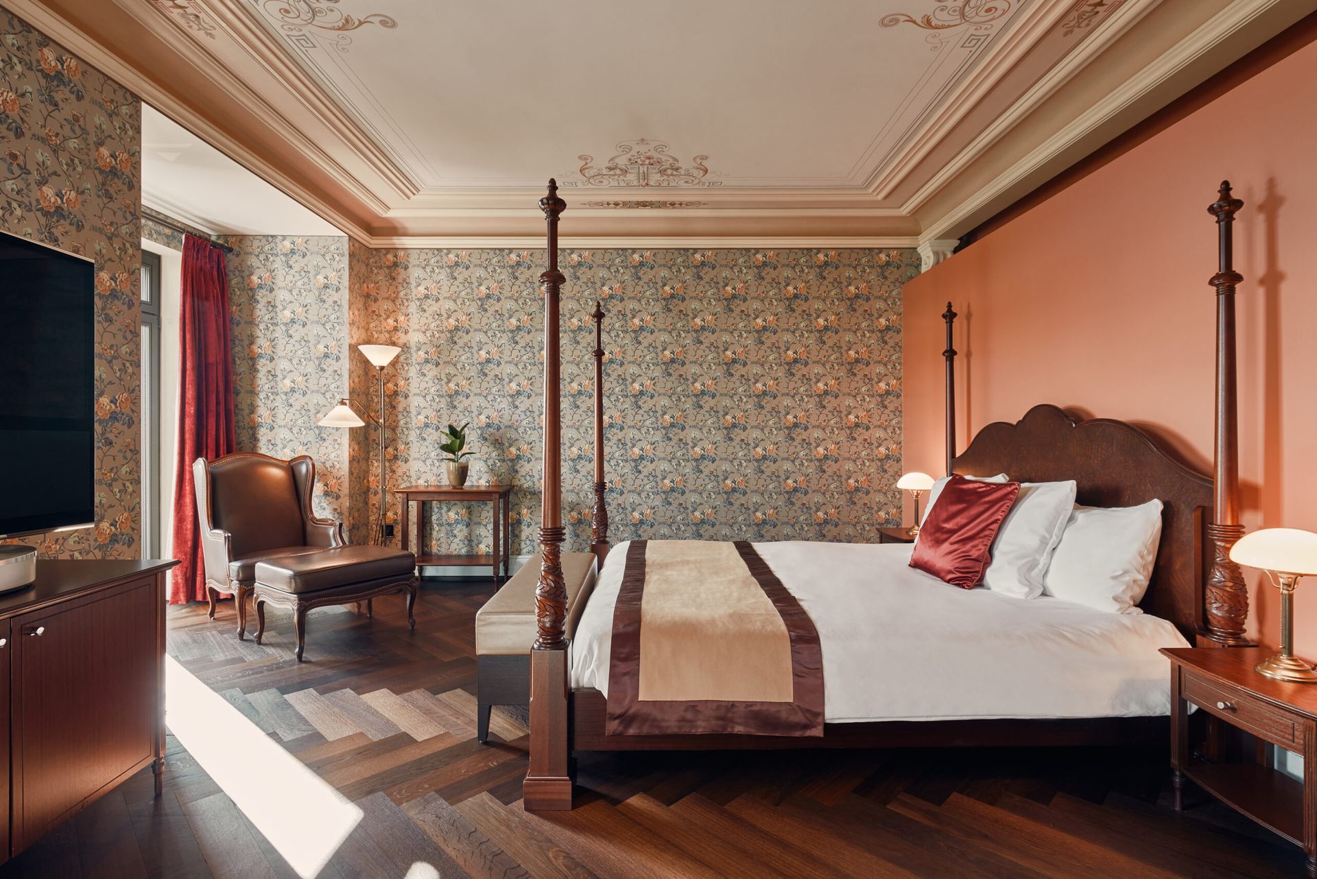 Elegant bedroom at this fairytale hotel in Zurich features a four-poster bed, floral wallpaper, peach accent wall, wooden floors, leather armchair, vintage desk, and large window with red curtains that let in natural light—true Dolder Grand charm.