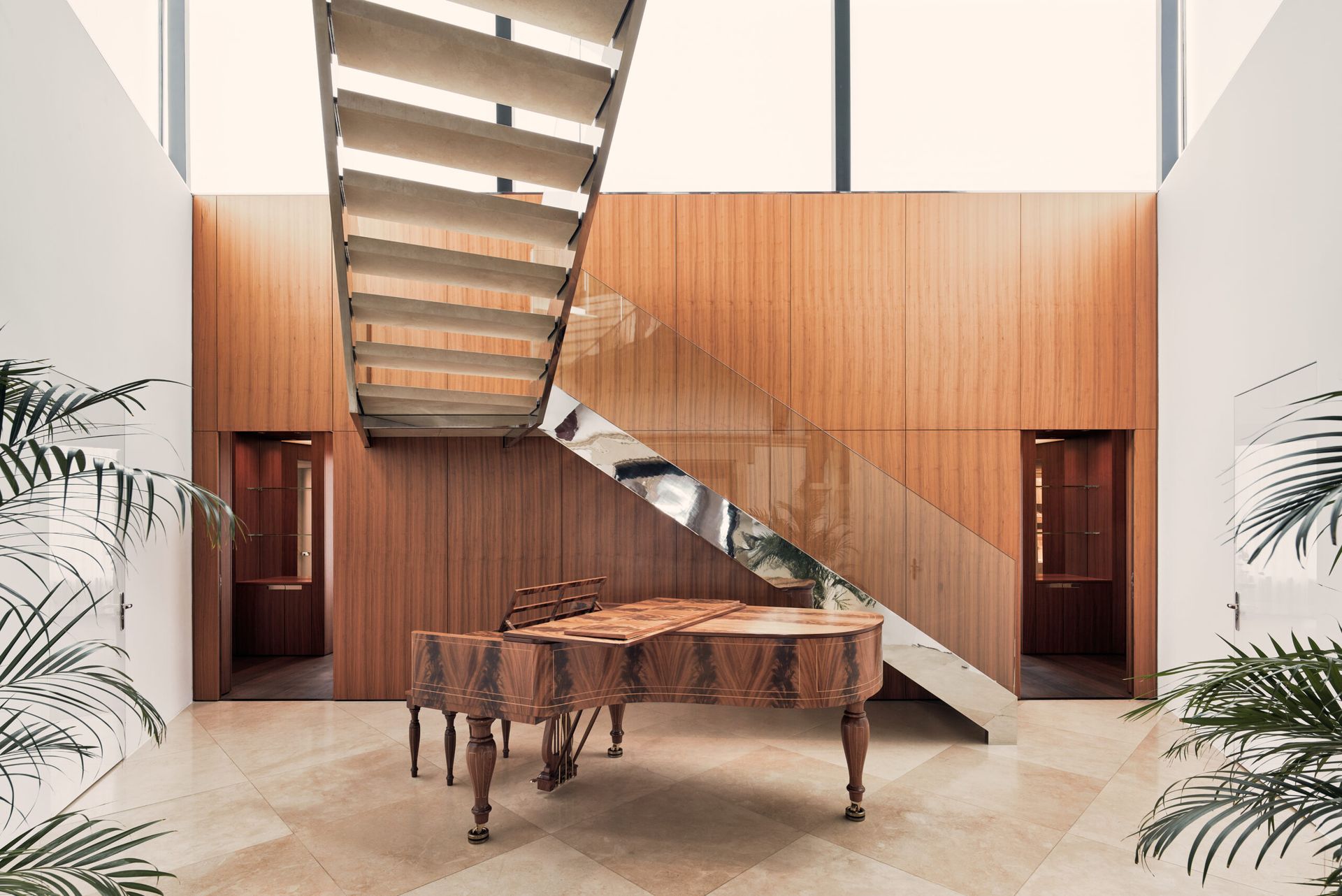 A grand piano sits on a beige marble floor beneath a modern floating staircase with a mirrored balustrade in a spacious, light-filled room with wooden wall panelling and tall windows at Zurich’s fairytale hotel, the Dolder Grand.