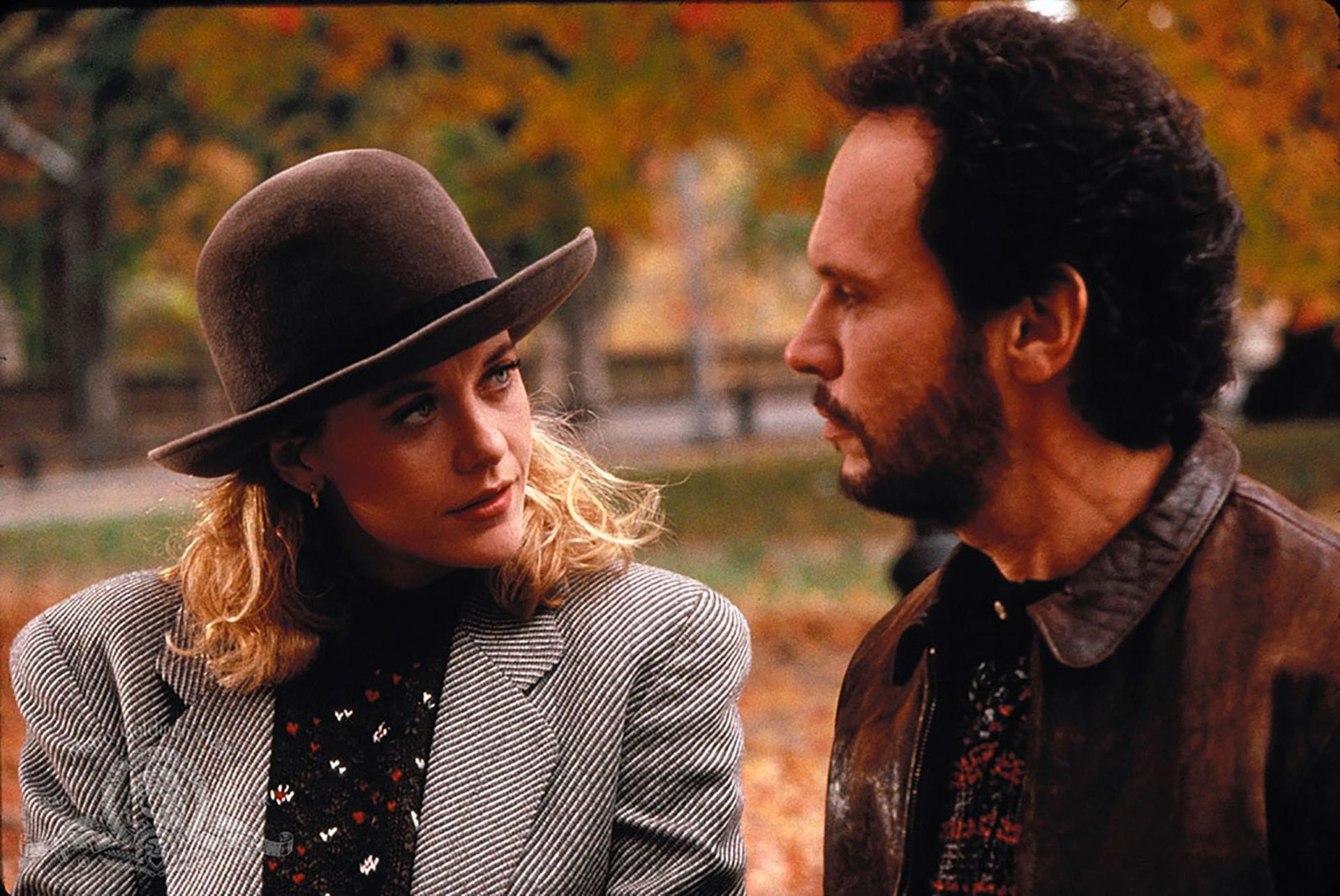 A woman in a grey blazer and brown hat looks at a man in a brown leather jacket as they talk outdoors, autumn leaves swirling around them—just the kind of scene you’d see from a bloke’s perspective in one of the best romantic comedies.