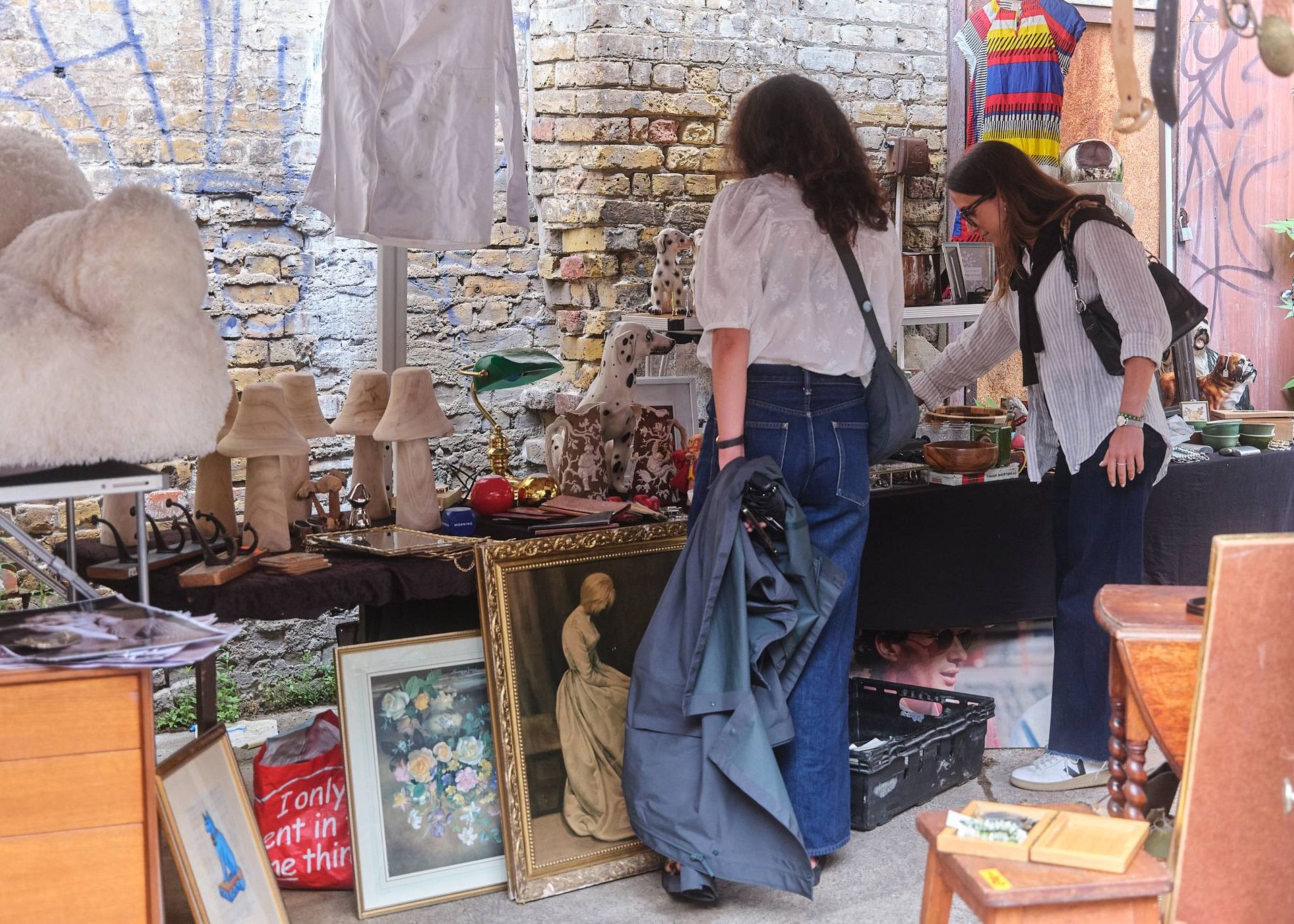 Two women browse a flea market stall filled with vintage items—framed paintings, lamps, ceramics, and soft toys. Set against a graffiti-covered brick wall, this scene captures the eclectic vibe of flea markets London is known for.