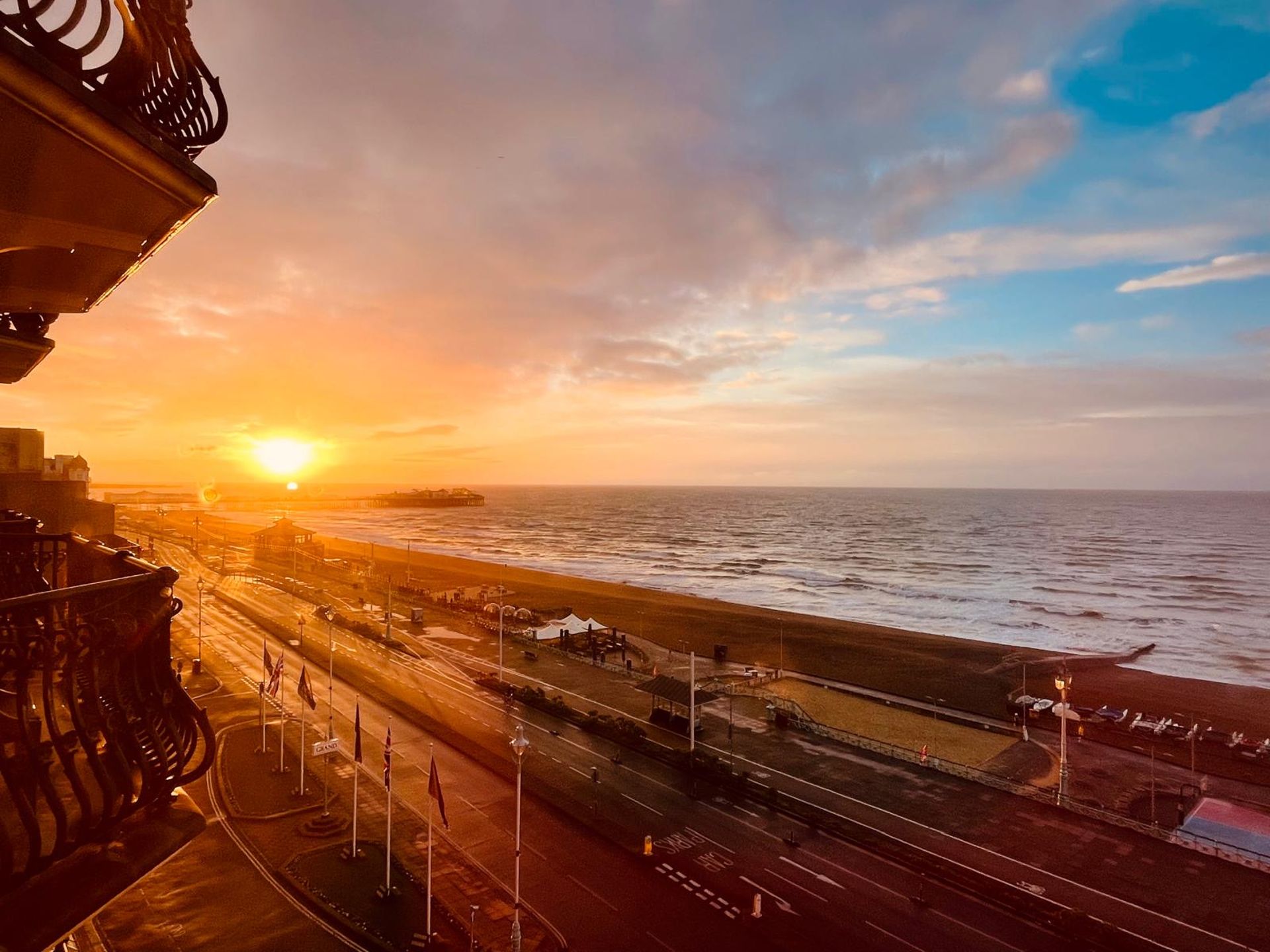 A vibrant sunrise over a beach and sea in Brighton, viewed from a balcony—one of the best things to do in Brighton. The sky features golden and blue tones, with a road lined with streetlights and flags running alongside the seafront.