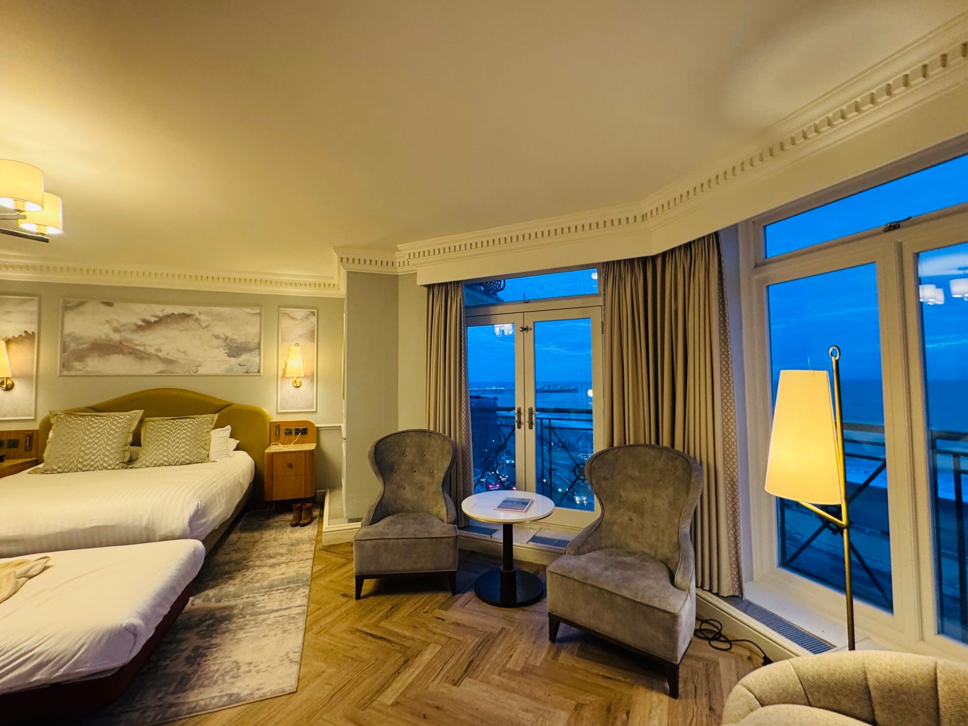 A hotel room with two beds, two armchairs around a small table by large windows, and a view of the sea and balcony at dusk. After exploring the best things to do in Brighton, relax in warm lighting and elegant furnishings.