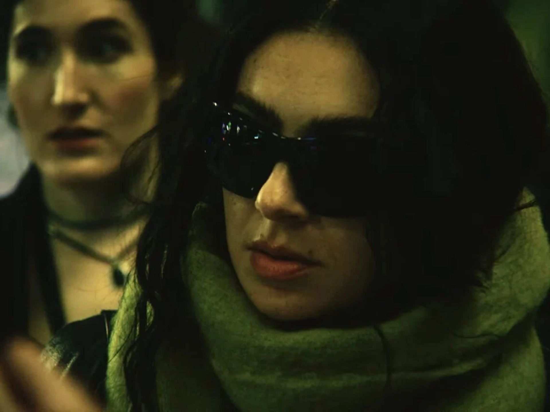 A woman with long dark hair wears large dark sunglasses and a thick scarf, looking serious—like a scene from The Moment, the new film. Another woman with pale skin and dark hair stands blurred in the background amid dim lighting.