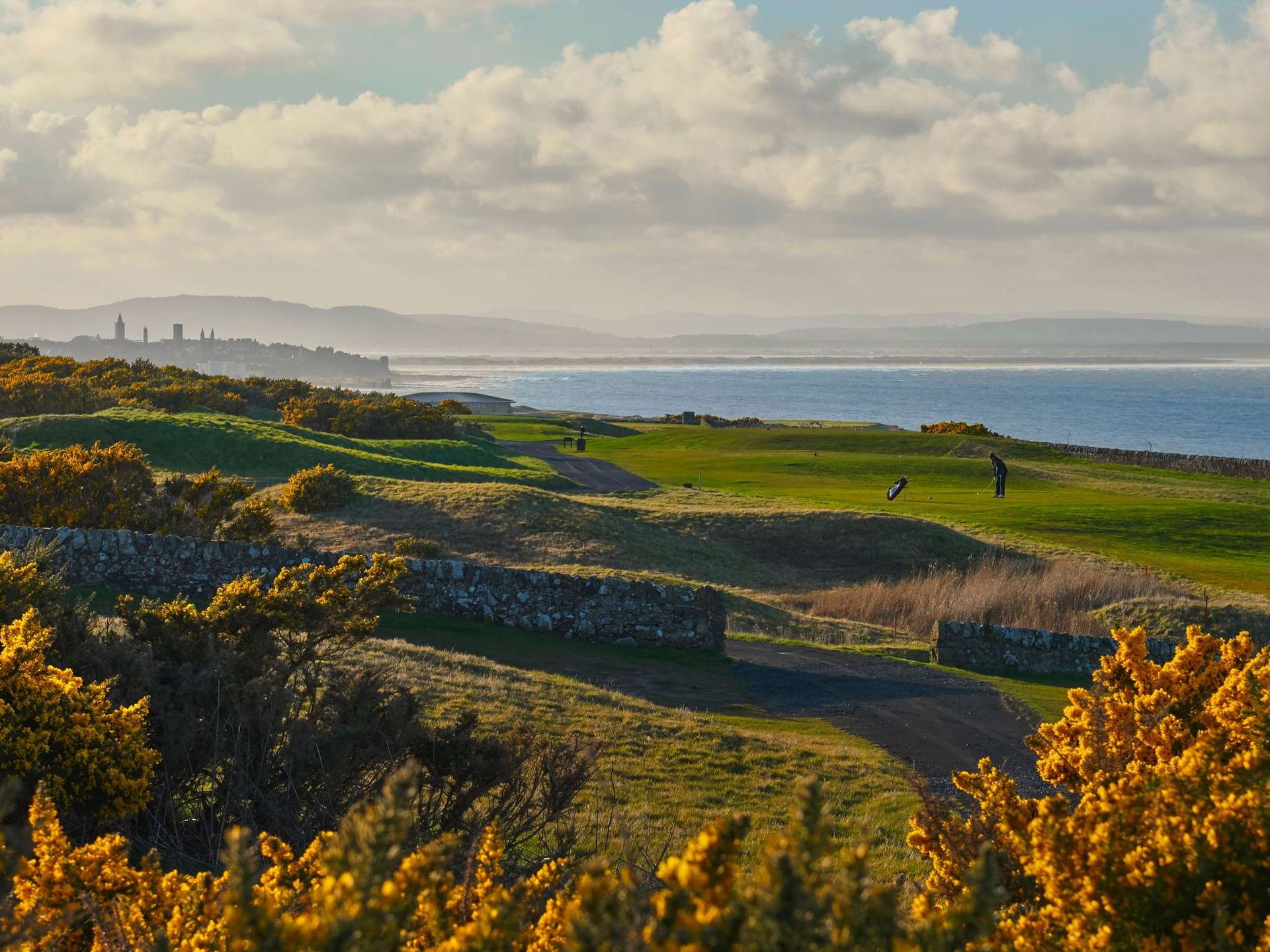 A scenic golf course near the sea with lush green fairways, a stone wall, yellow flowering bushes, and distant hills under a partly cloudy sky. A few golfers enjoy the course, ideal for guests seeking UK Golf Hotels nearby.