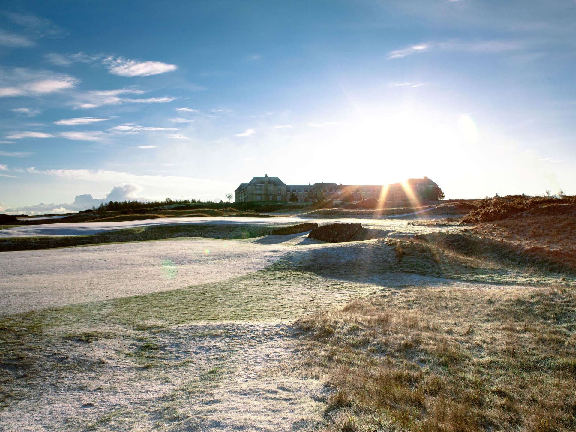 A frosty golf course under a clear blue sky with the sun rising behind a large building—reminiscent of luxury hotels in Scotland—casting long shadows and creating a serene morning scene.