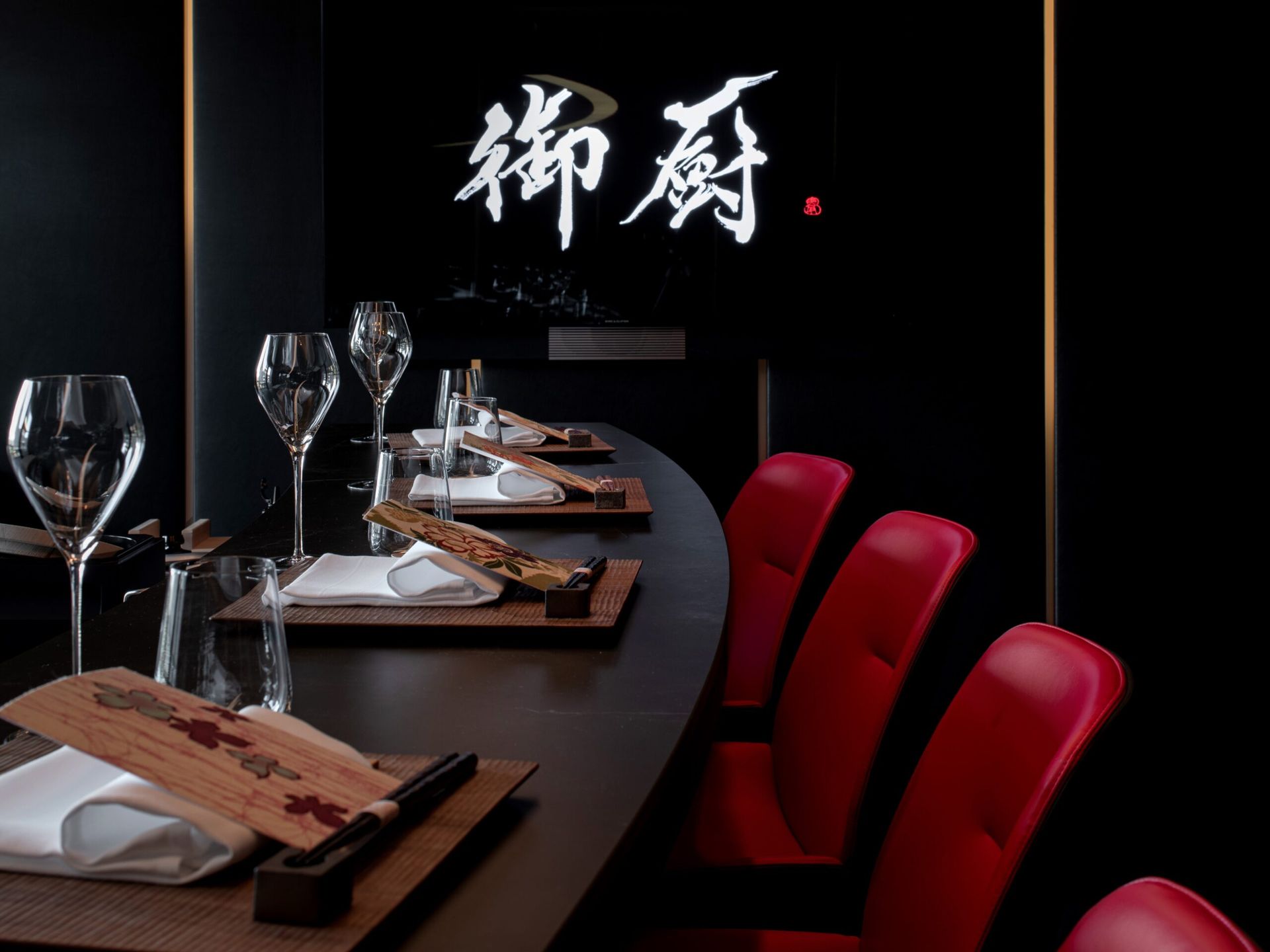 A sleek restaurant table set with wine glasses, plates, and wooden chopsticks on place mats, with red chairs and illuminated Japanese kanji characters on a black wall—evoking the refined ambience found at Zurich's Dolder Grand fairytale hotel.