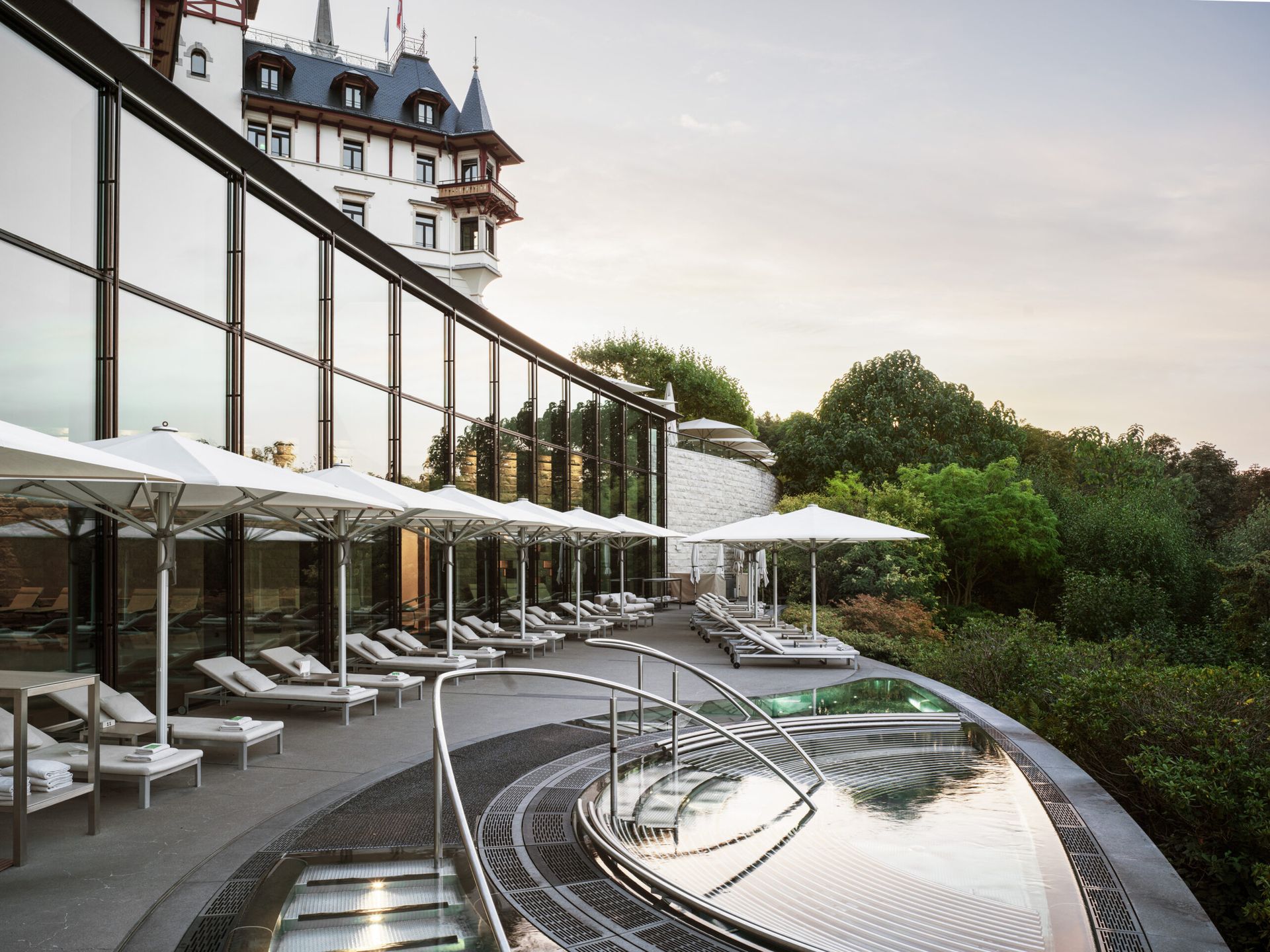 A modern outdoor swimming pool area with loungers and white parasols, set beside a building with large glass windows, overlooks the Dolder Grand—a fairytale hotel in Zurich—framed by lush green trees and a historic castle-like structure in the background.