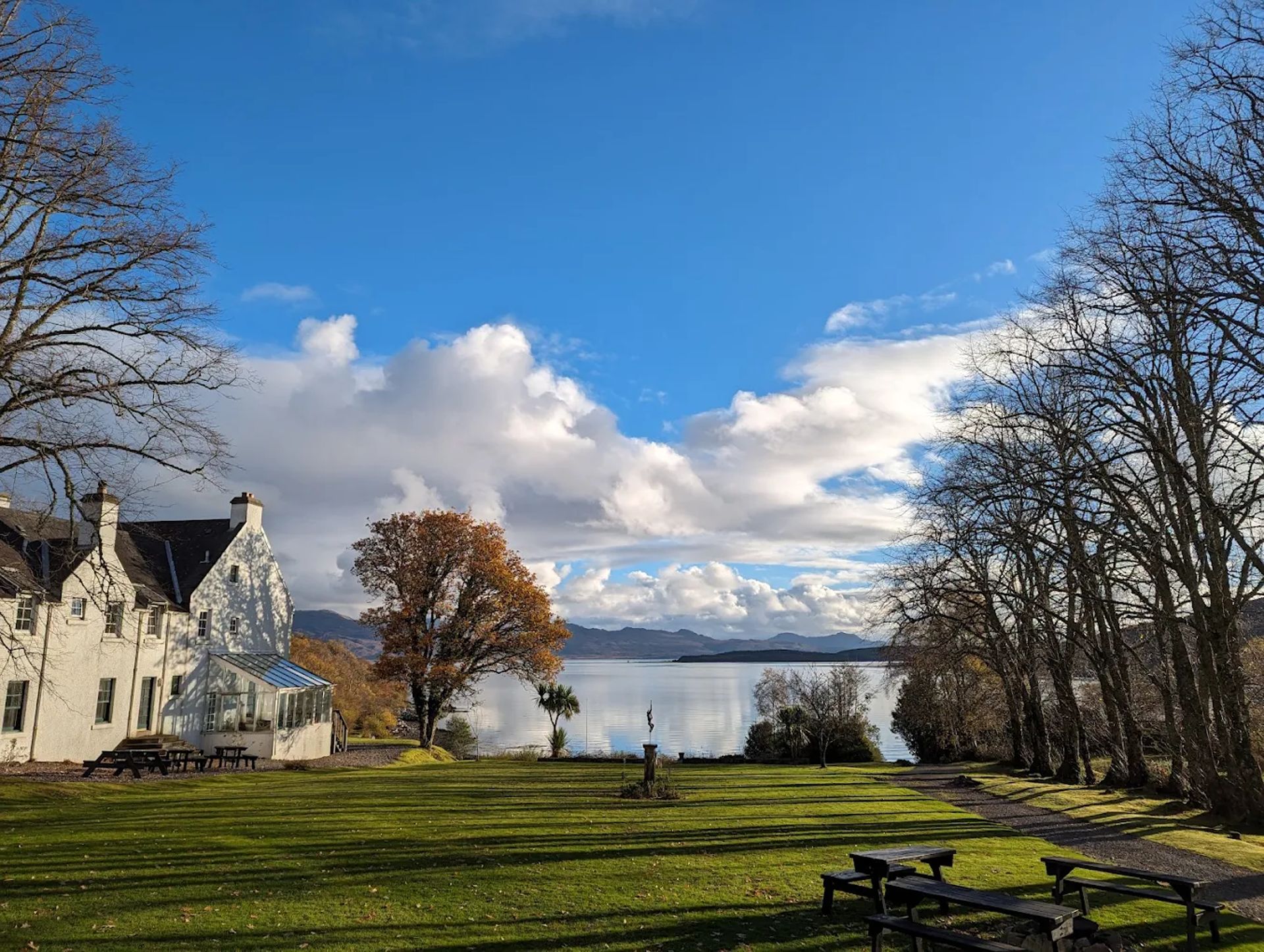 A large white house, possibly Kinloch Lodge, stands next to a lawn with benches, bordered by leafless trees. Beyond the lawn, a calm lake reflects the blue sky and clouds, with hills visible in the distance.