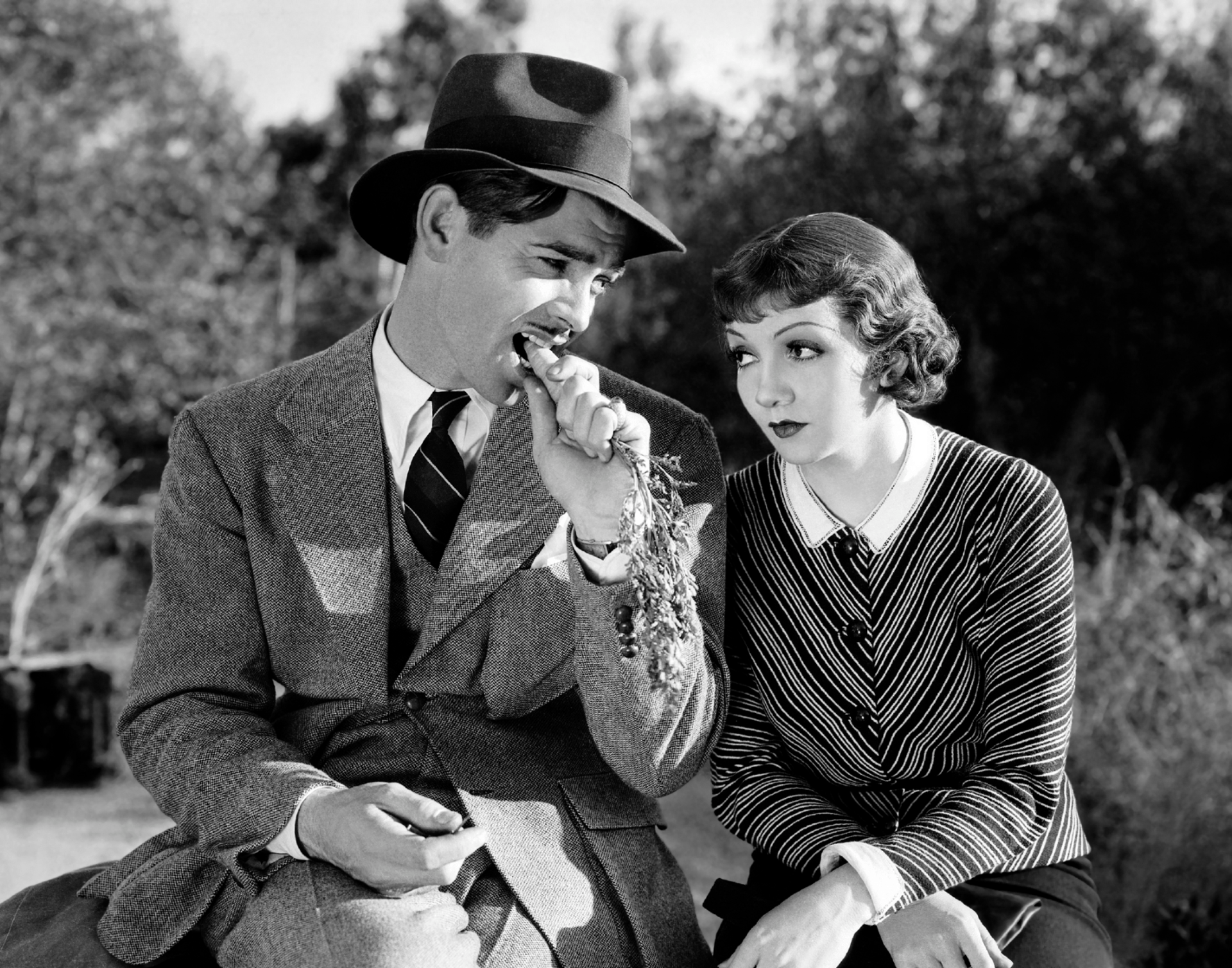 A man in a suit and hat sits outdoors, eating a carrot as a woman beside him, dressed in stripes, looks on with curiosity—the scene echoes quirky moments often found in the best romantic comedies from the bloke's perspective.