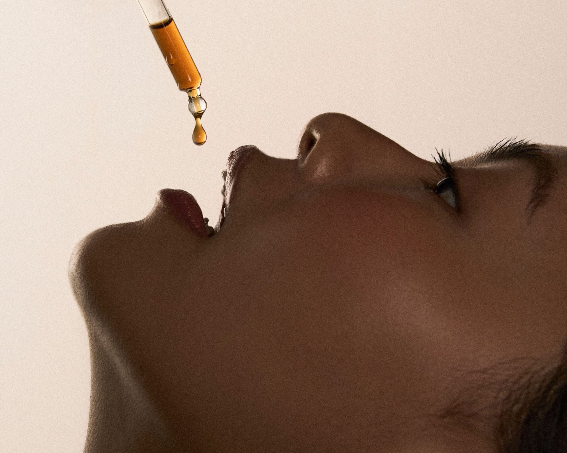 A close-up side view of a person’s face with their mouth open as a dropper dispenses amber-coloured liquid towards their lips—a scene that captures the essence of London Cool Girls who have it all. The background is neutral and uncluttered.