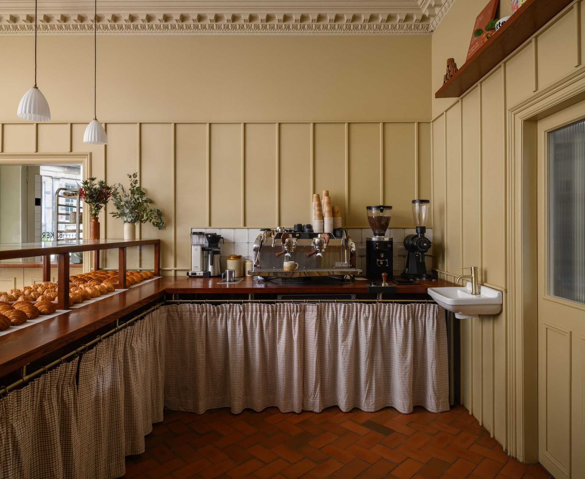 A cosy bakery kitchen inspired by Edinburgh, complete with a coffee station, pastries on display, a sink, and chequered curtains beneath the counter. The cream-coloured walls with panelling and warm hanging lights invite you into the space—an insider’s guide to charming travel destinations brought to life through cosy details.