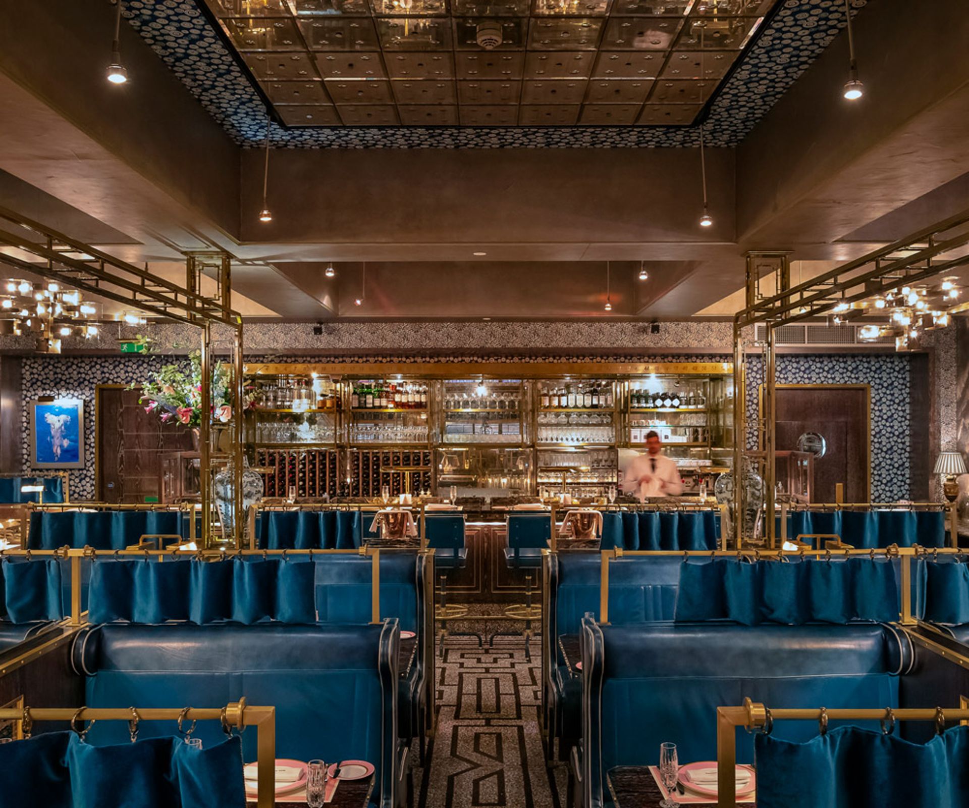 A luxurious restaurant interior with plush blue velvet banquettes, gold accents, patterned walls, and a mirrored ceiling. Curated by Hannah Crosbie, the bartender stands behind a well-stocked bar amid warm lighting and elegant decor.