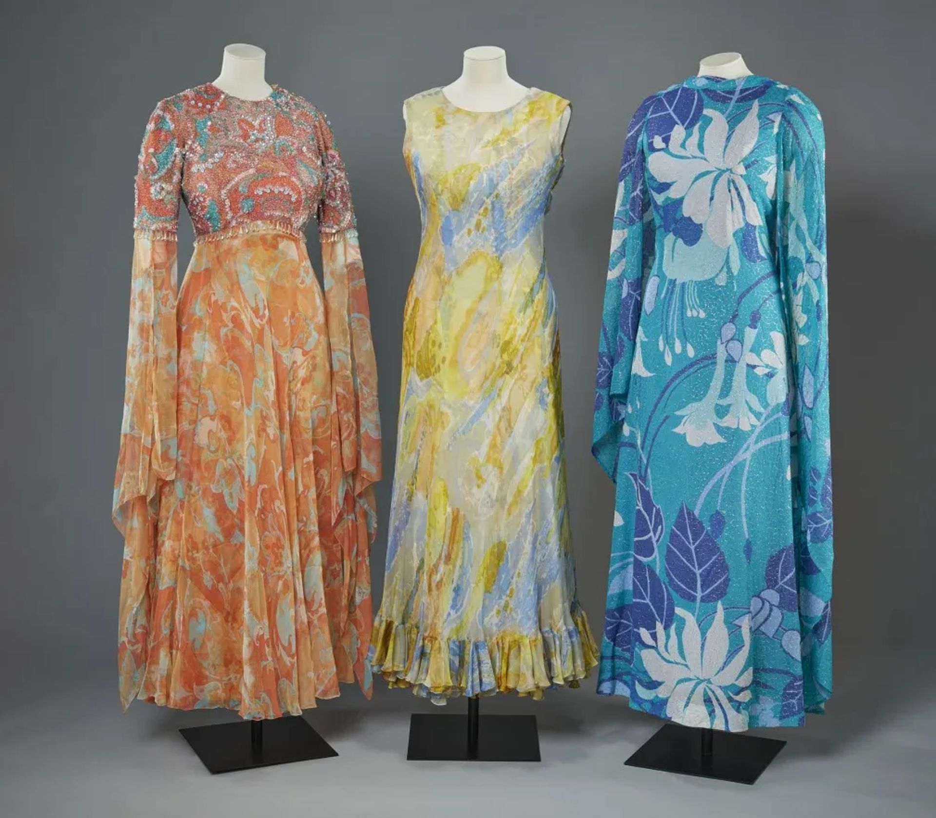 Three vintage mannequins display 1960s-style dresses, perfect for Fashion Exhibitions: a long orange floral gown with bell sleeves, a sleeveless yellow and blue watercolour dress, and a blue and purple floral dress with a matching cape.