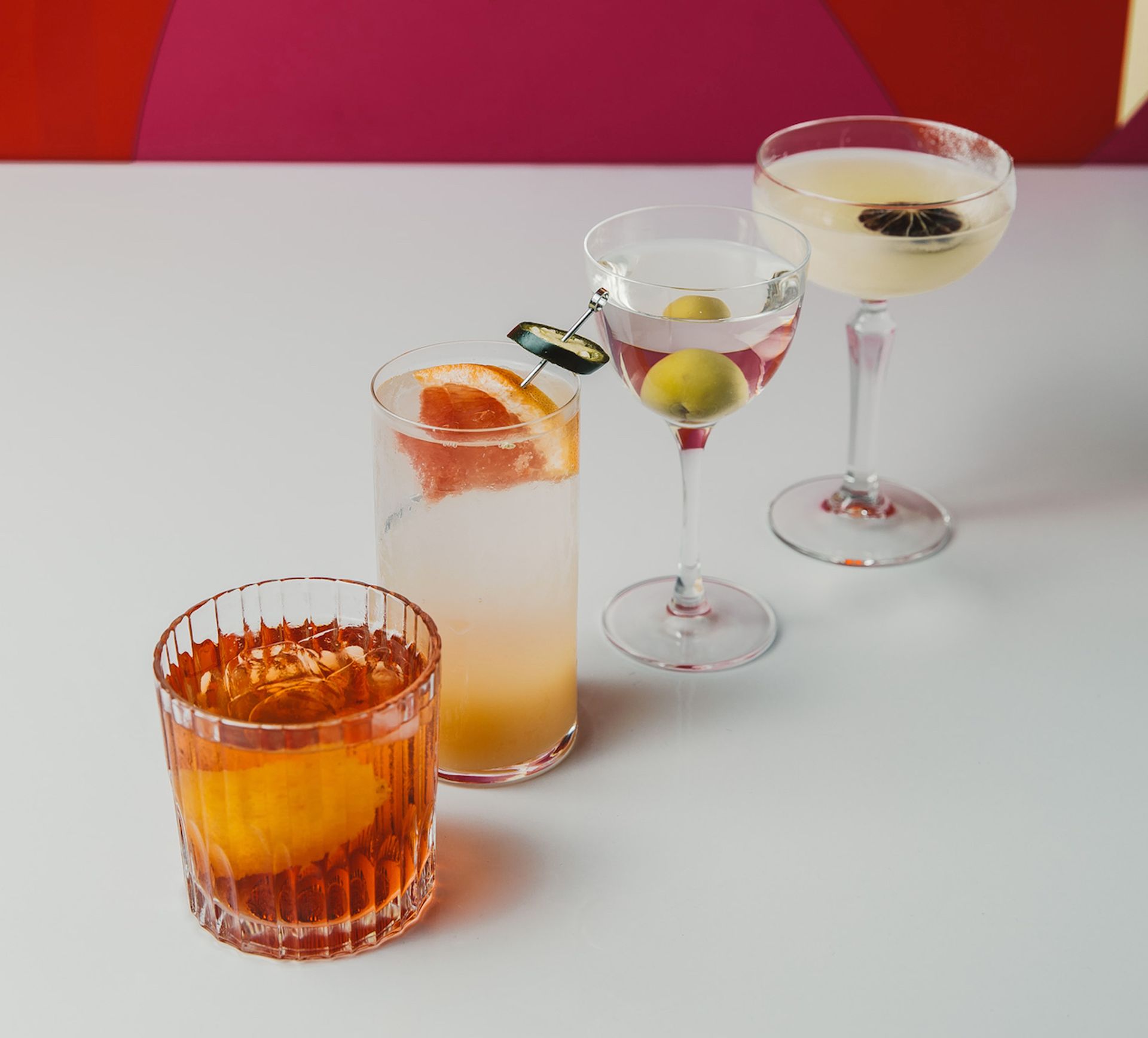 Four cocktails on a white table: an orange drink with an orange slice, a tall grapefruit cocktail, a martini with olives, and a Margarita garnished with dried citrus. Perfect for any event, set against a bold, colourful background.