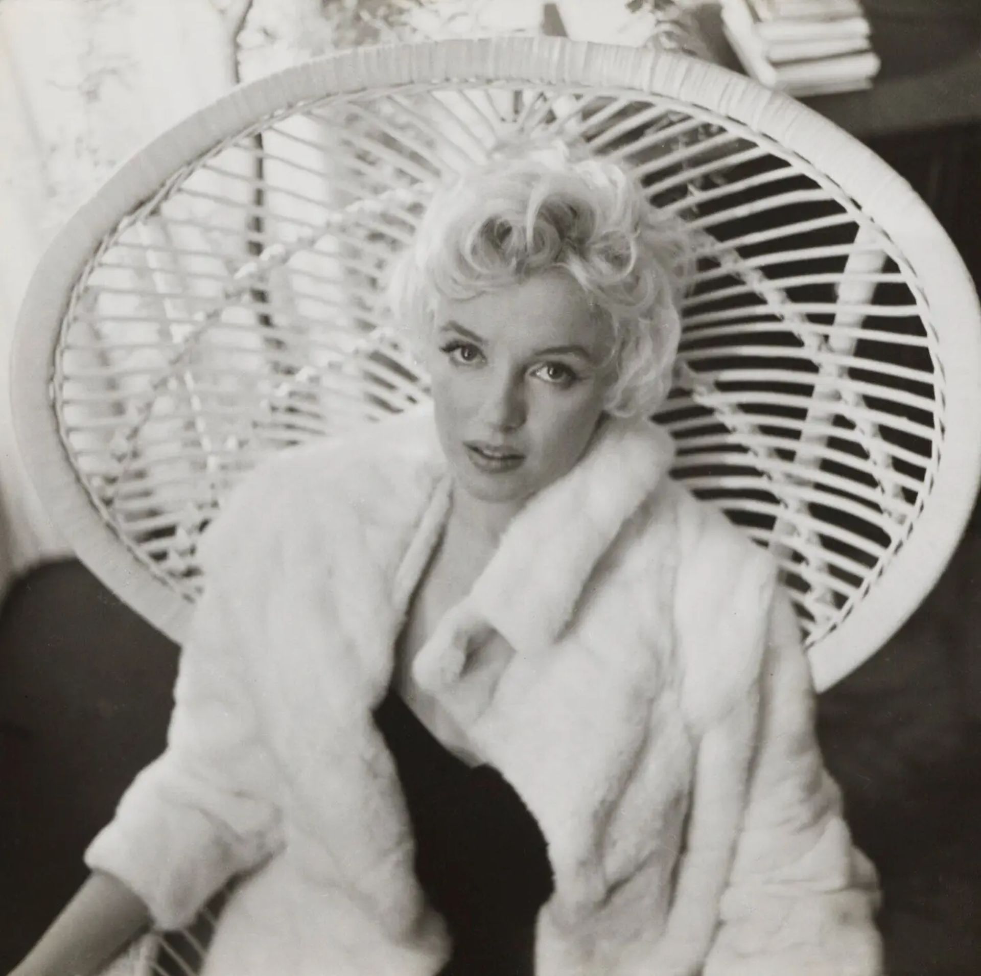 A woman with short curly blond hair sits in a large, circular wicker chair, wearing a light-coloured fur coat over a dark outfit, looking up slightly with a calm expression—ready for the season’s top Fashion Events.