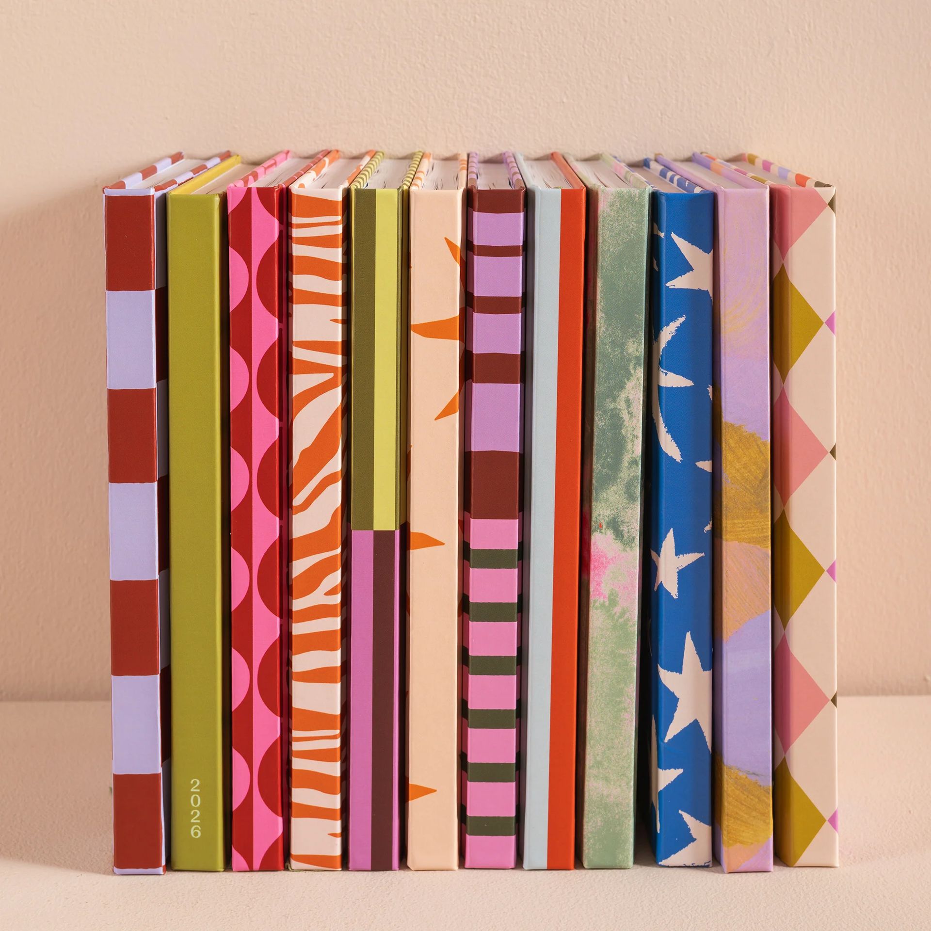 A row of colourful 2026 diaries featuring vibrant patterns—stripes, zigzags, polka dots, stars, and abstract designs—stands upright against a light background.