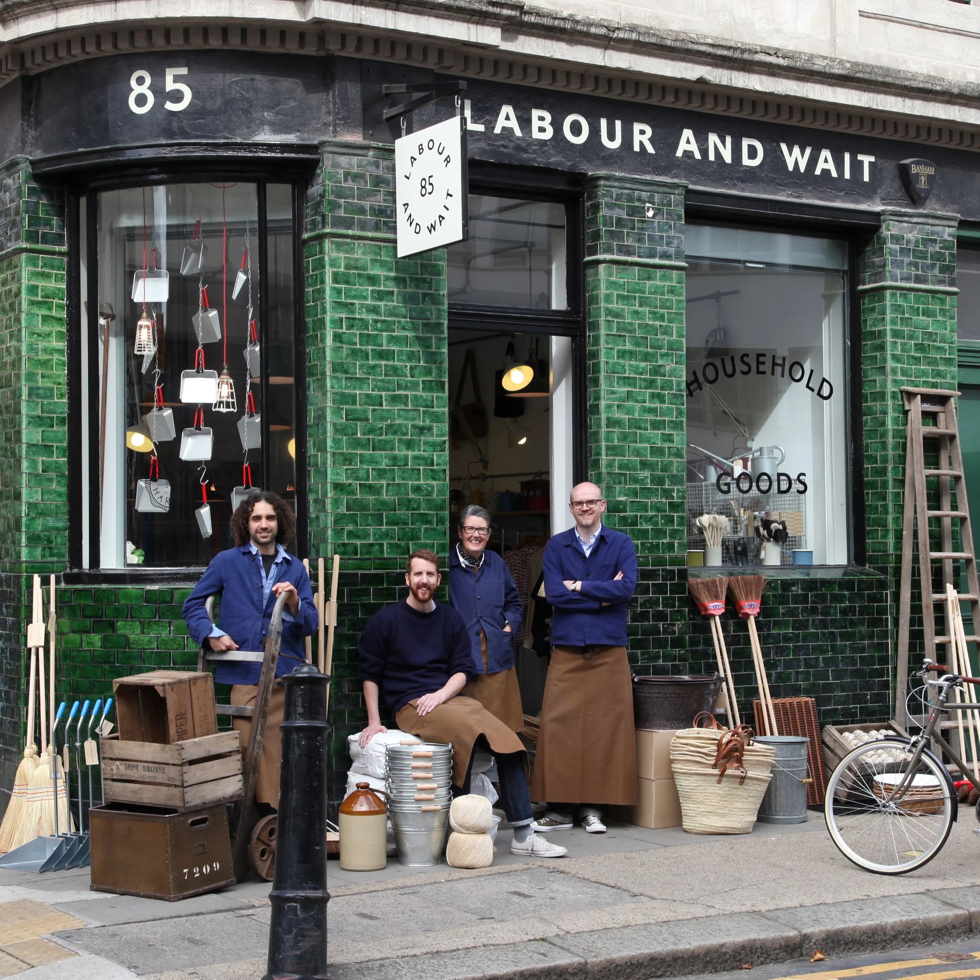 Four people in blue shirts and brown aprons stand and sit outside Labour and Wait, a shop with a green-tiled shopfront displaying household goods like brushes, baskets, buckets, and office supplies. A bicycle is parked nearby.