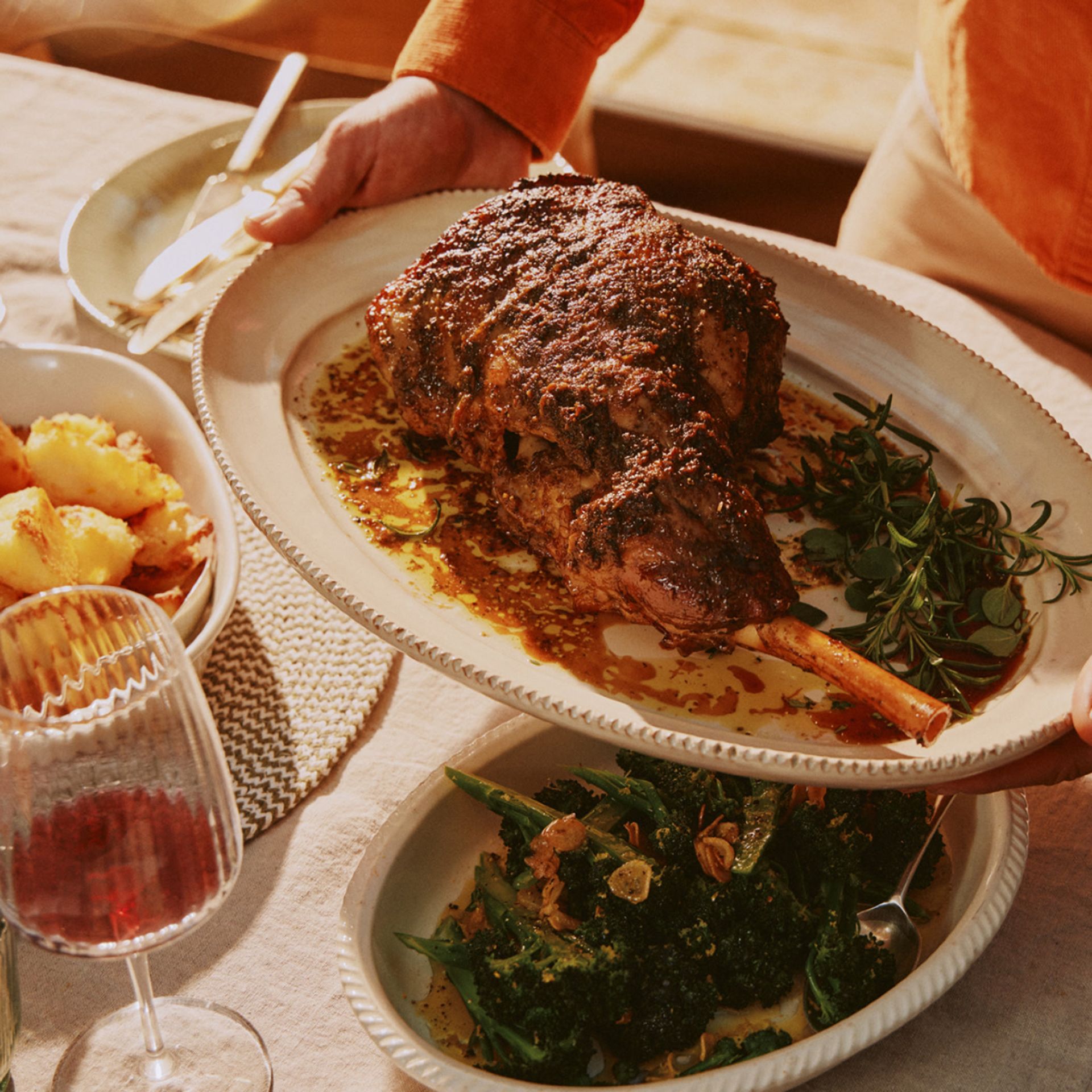 A person serves a platter of roast lamb garnished with herbs—perfect for Easter entertaining. Nearby are dishes of roast potatoes, sautéed greens, and a glass of red wine on a beautifully laid dining table.