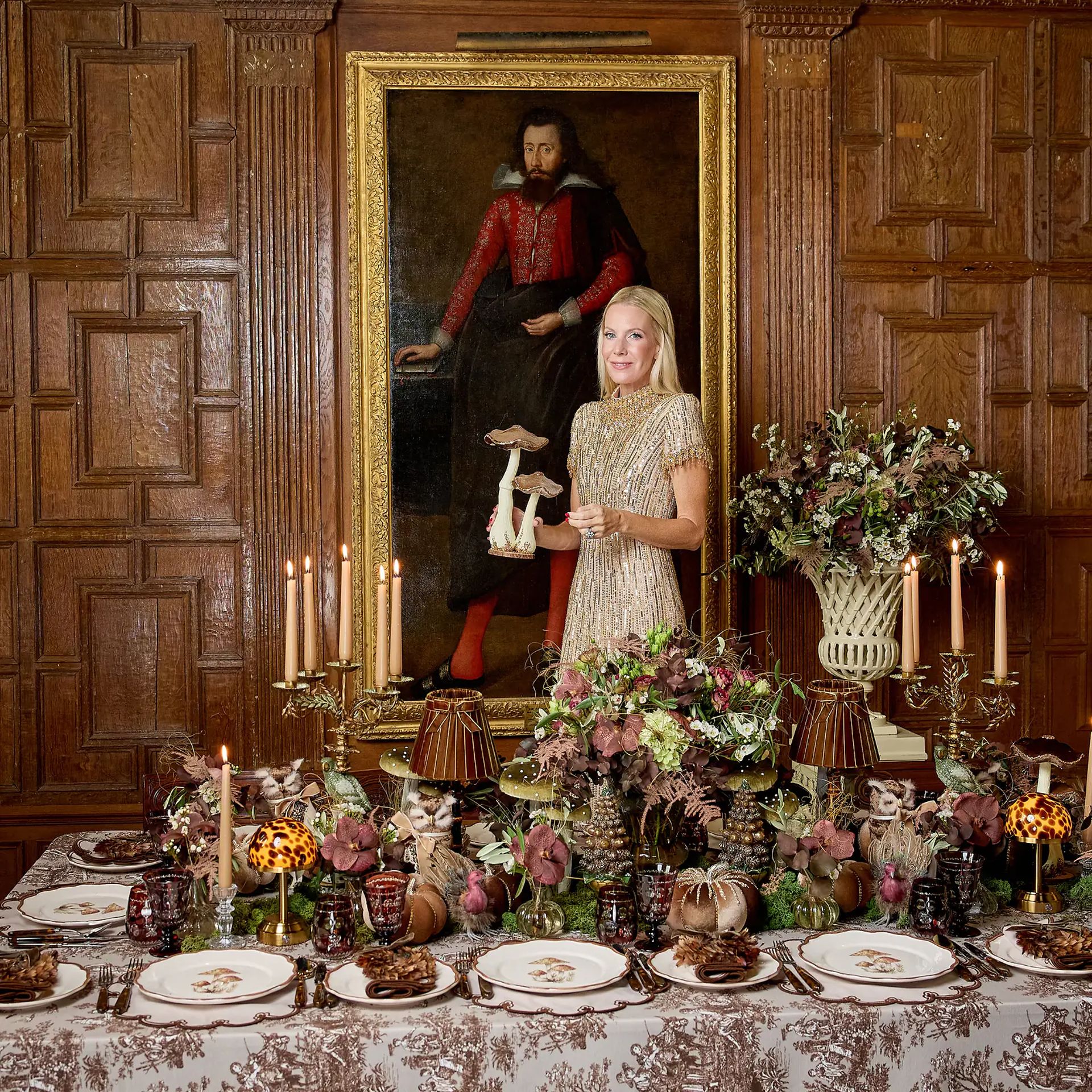 A woman in a beaded dress stands at an autumn tablescape, holding a decorative mushroom ornament in front of an oil painting of a man.