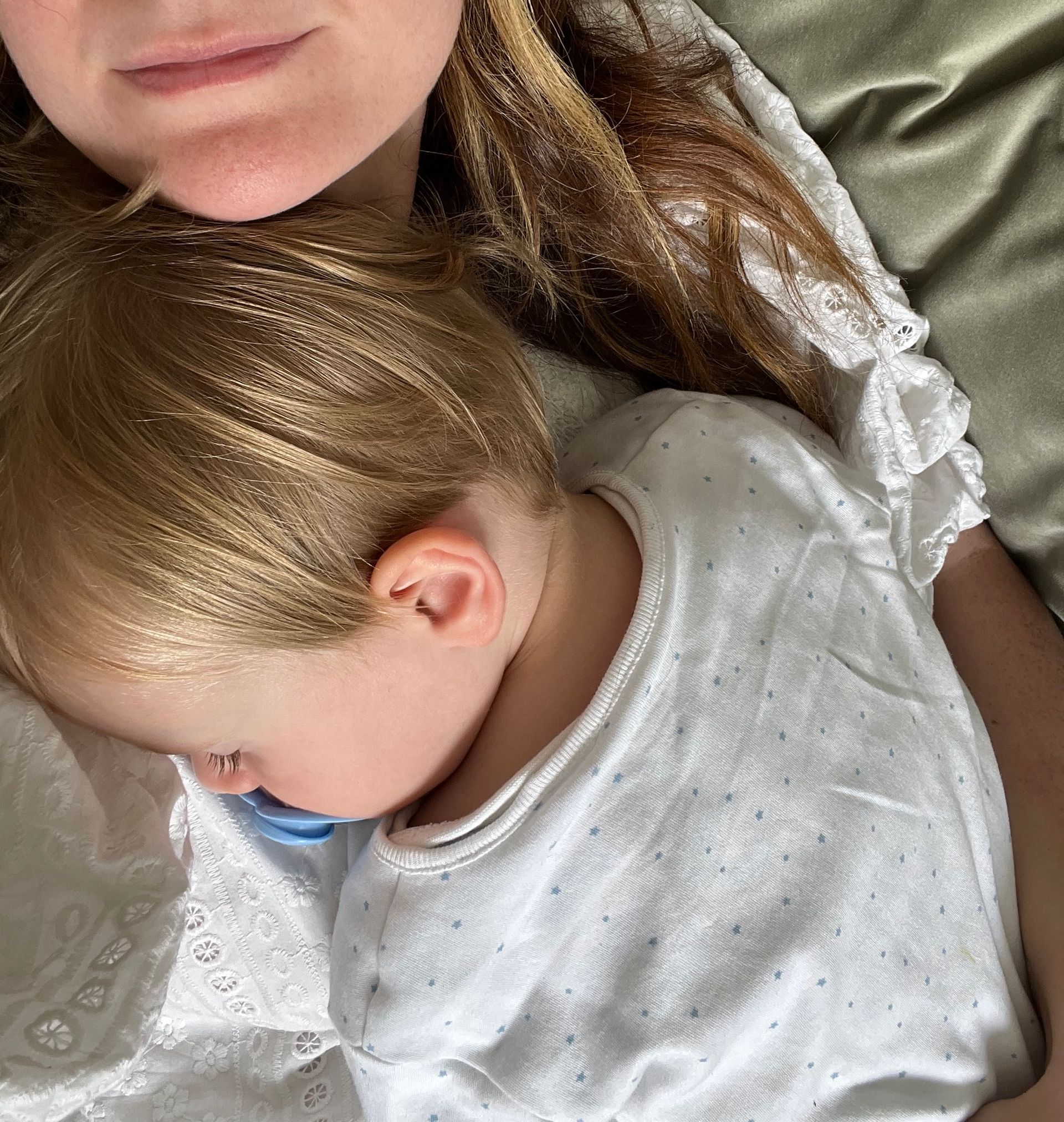 A baby with fair hair, wearing a white babygro, sleeps on a single mum’s chest. The adult, dressed in a white embroidered top, gently holds the baby while lying on a bed with light green bedding.