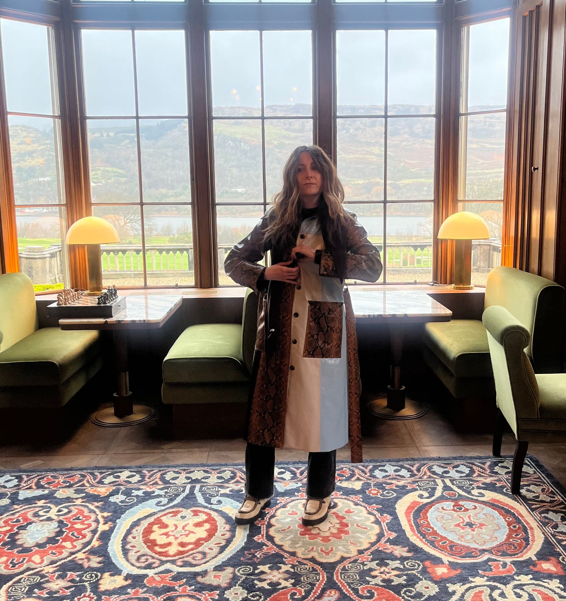 A woman with wavy hair stands in a room with large windows overlooking a lake. She wears a long, multi-textured coat featuring brown snakeskin and light blue panels—one of those signature pieces of “clothes my boyfriend hates.” She stands on a decorative rug between two small tables with green banquettes.
