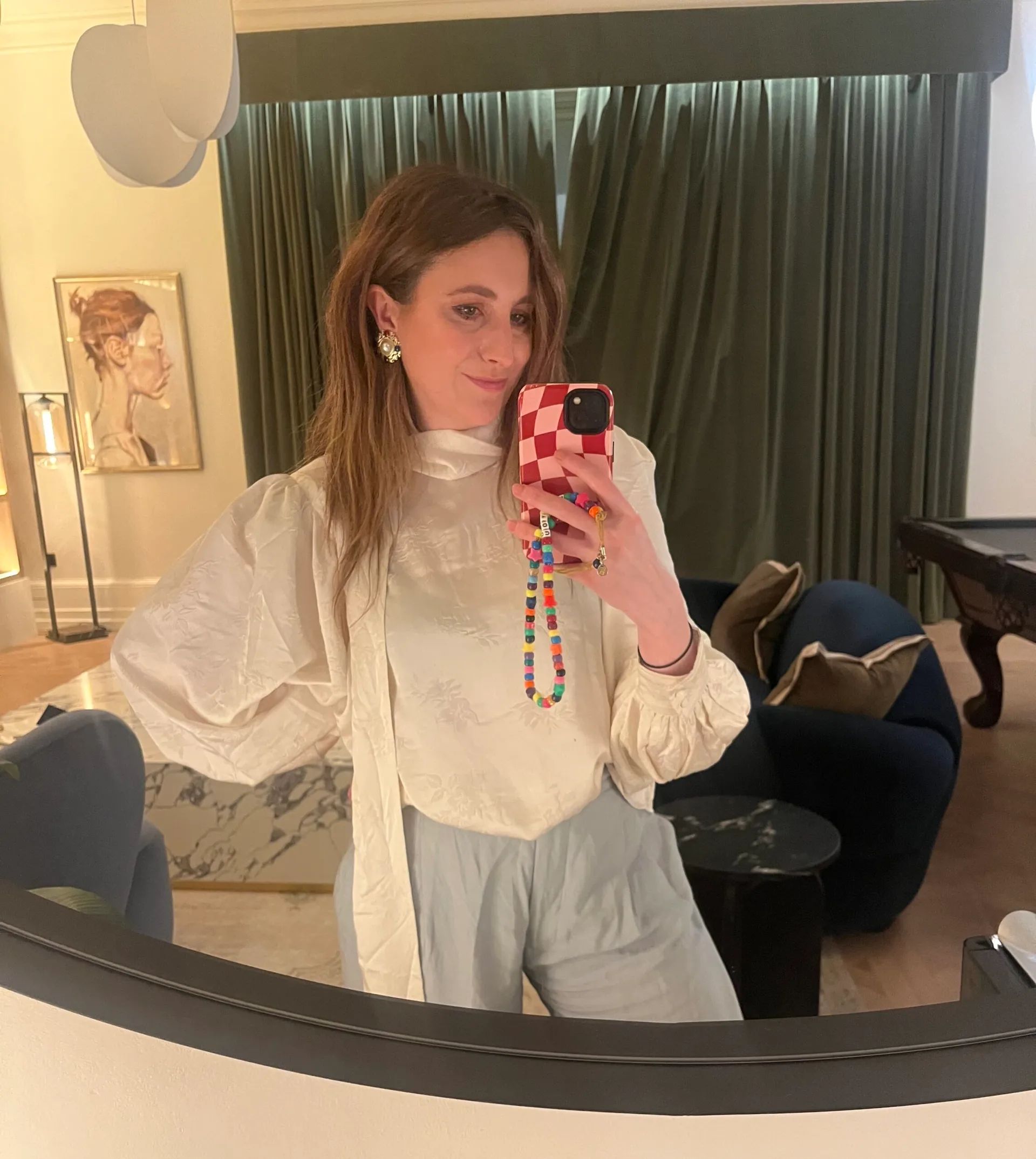 A woman with long brown hair takes a mirror selfie in a stylish room, showing off her fashion sense in a white blouse and light trousers. The background features green curtains, a blue chair, a marble table, and wall art.
