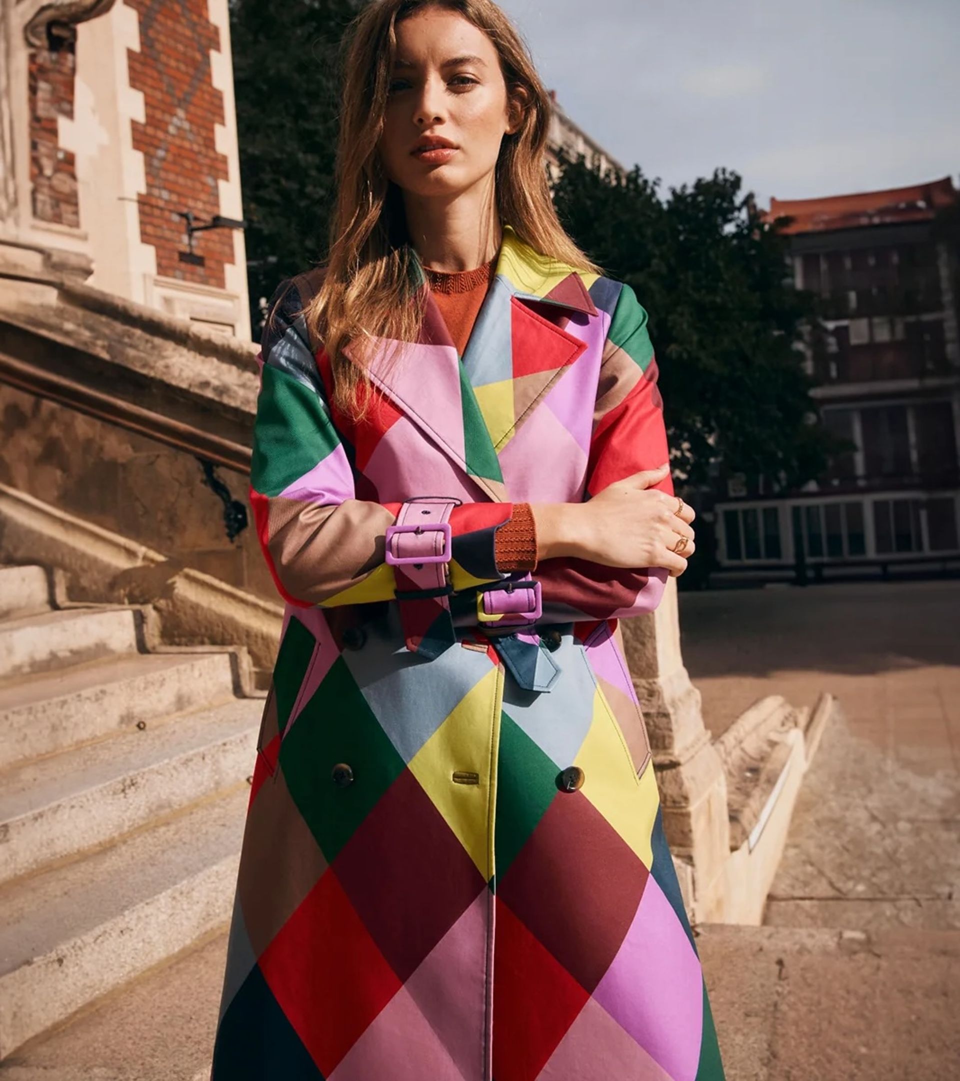 A woman stands outdoors on stone steps, wearing a colourful, diamond-patterned trench coat in bold Boden fashion shades of red, green, purple, yellow, and blue, with her arms crossed and a neutral expression.