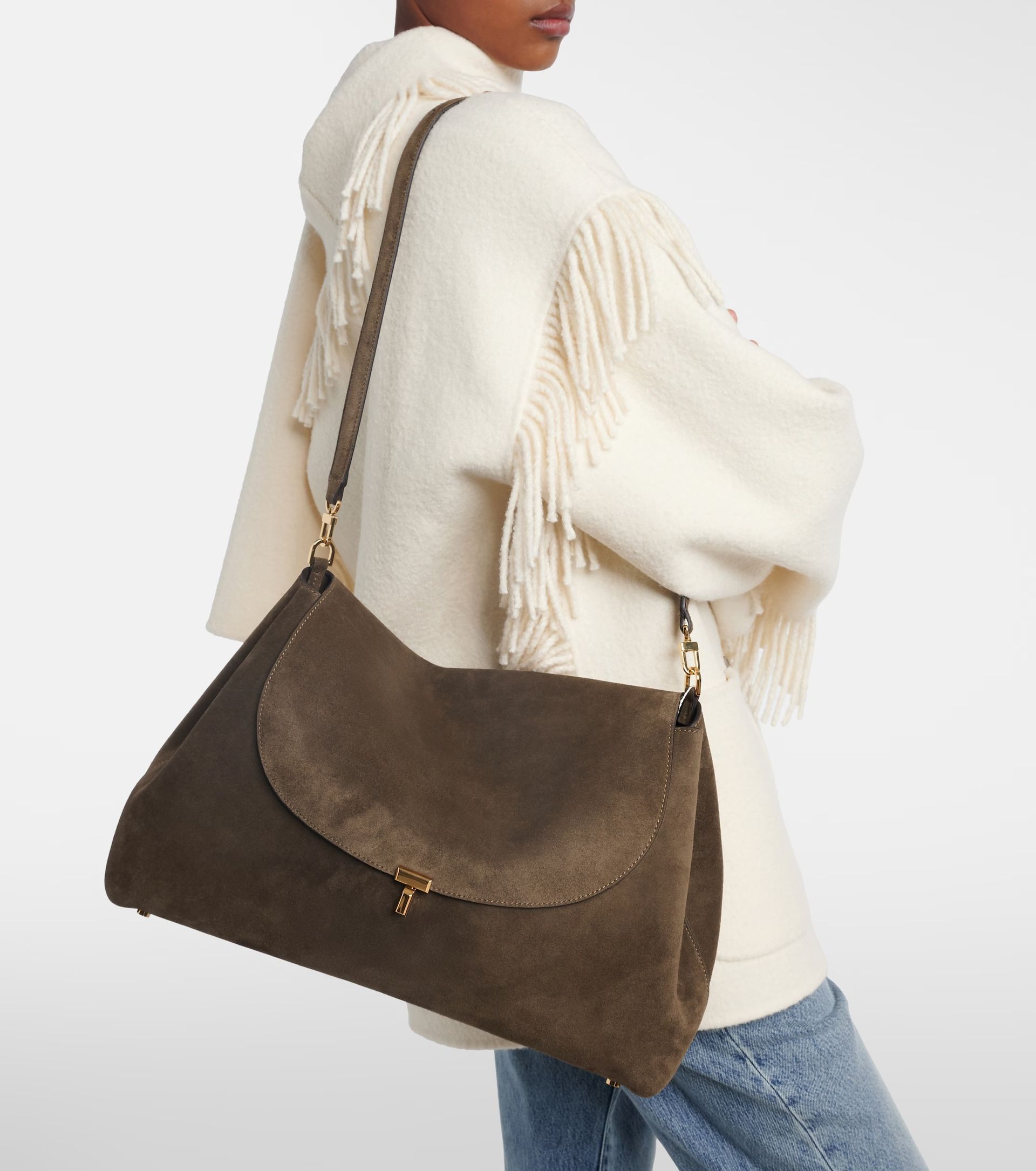 A person wearing a cream fringe coat and blue jeans holds a large brown suede shoulder bag with gold fittings—perfect for the summer sale season. The photo is cropped at the person’s nose.